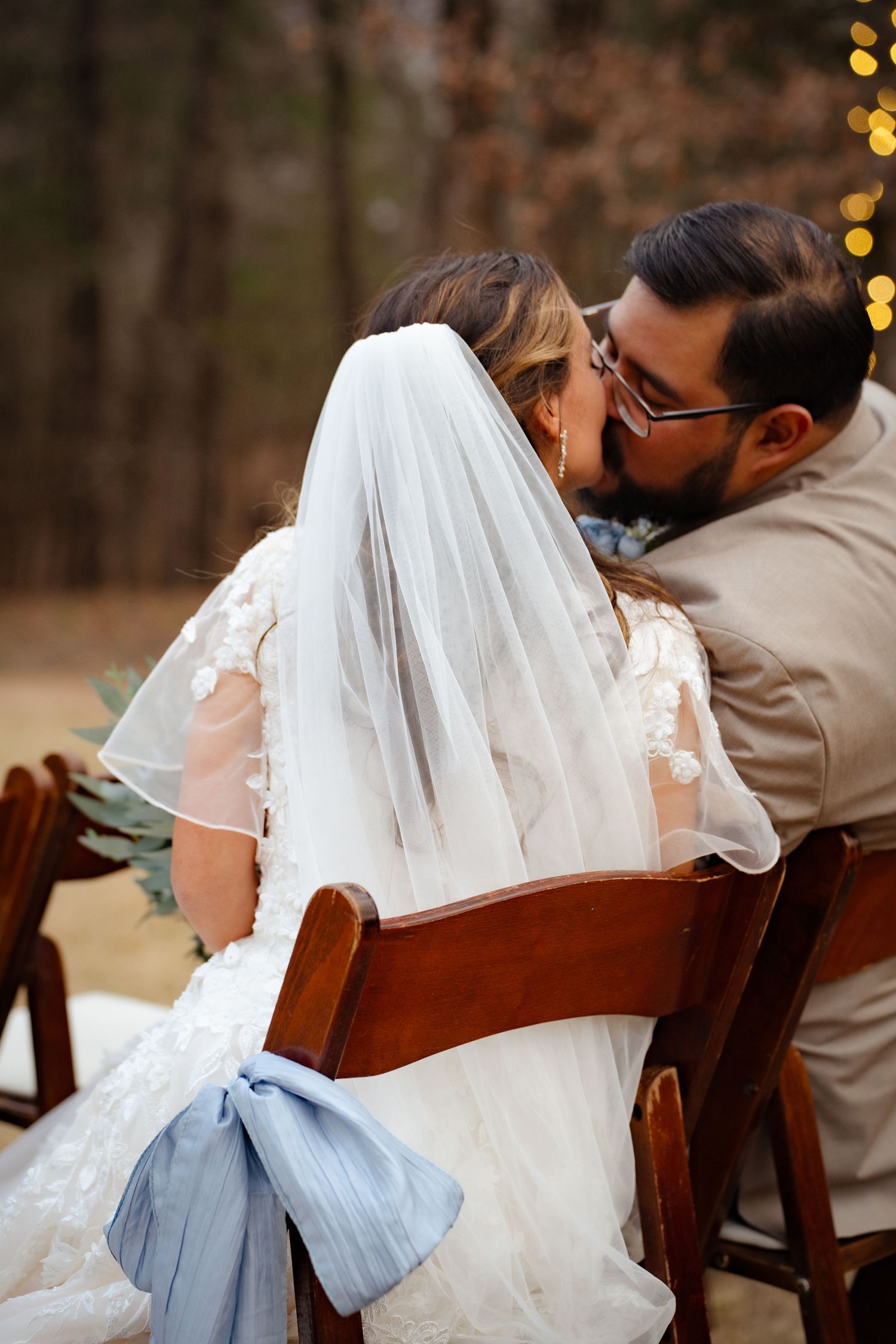 Bride and groom sharing a kiss while seated at outdoor wedding ceremony with blue ribbon decor.