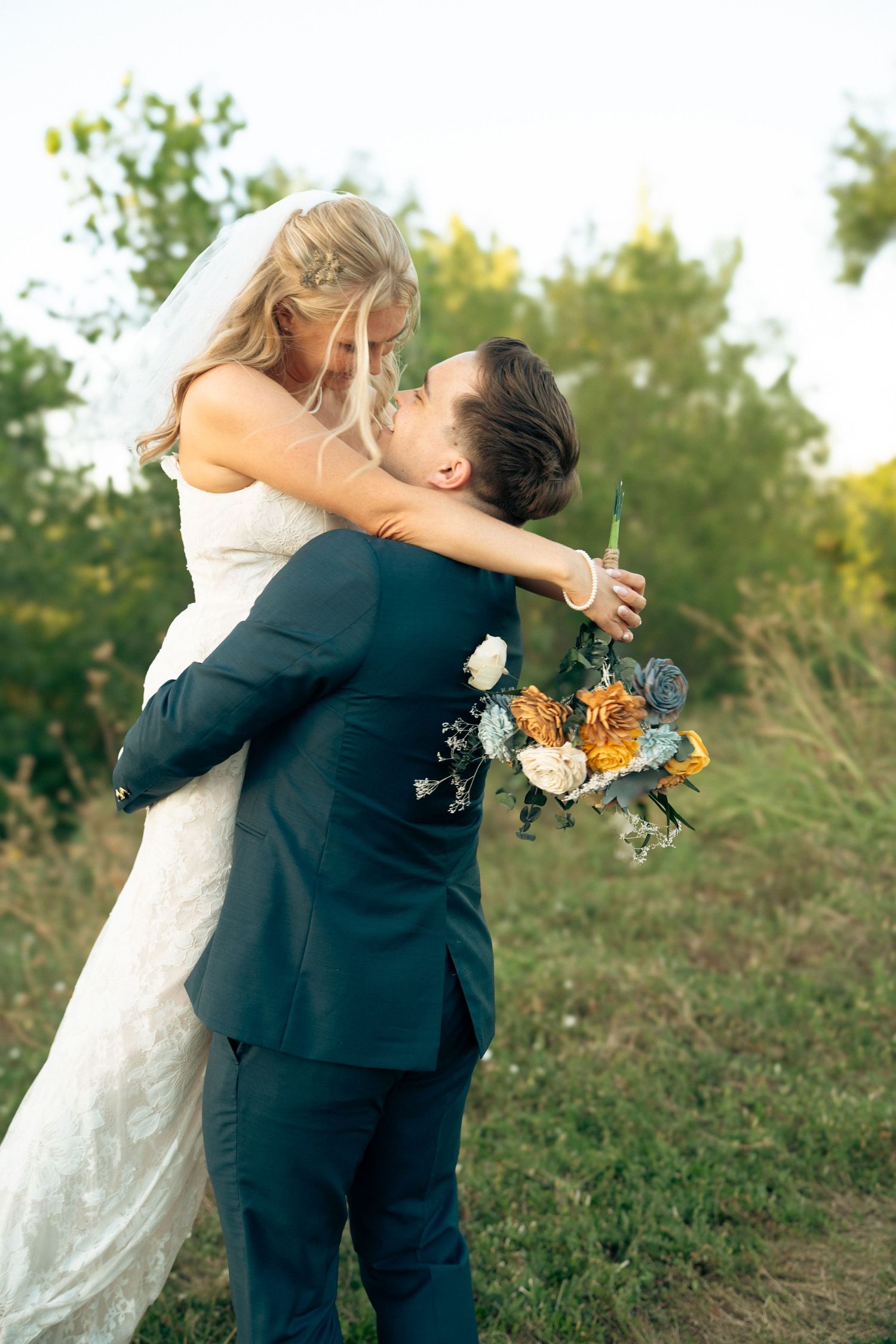 Groom lifts bride in white wedding dress while she holds colorful bouquet of orange and blue flowers.
