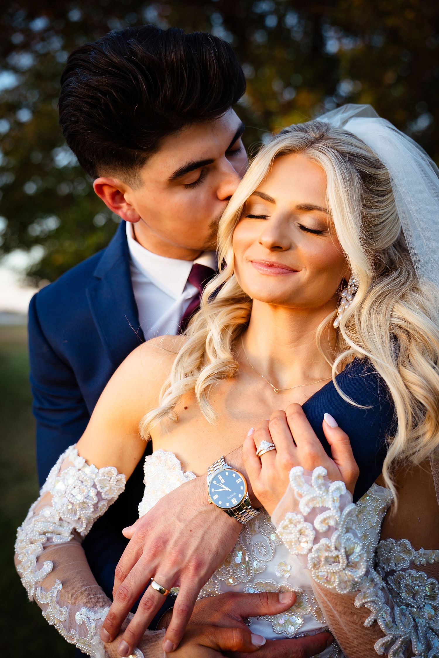 Groom tenderly kisses bride on the cheek as she smiles peacefully in golden autumn light outdoors.
