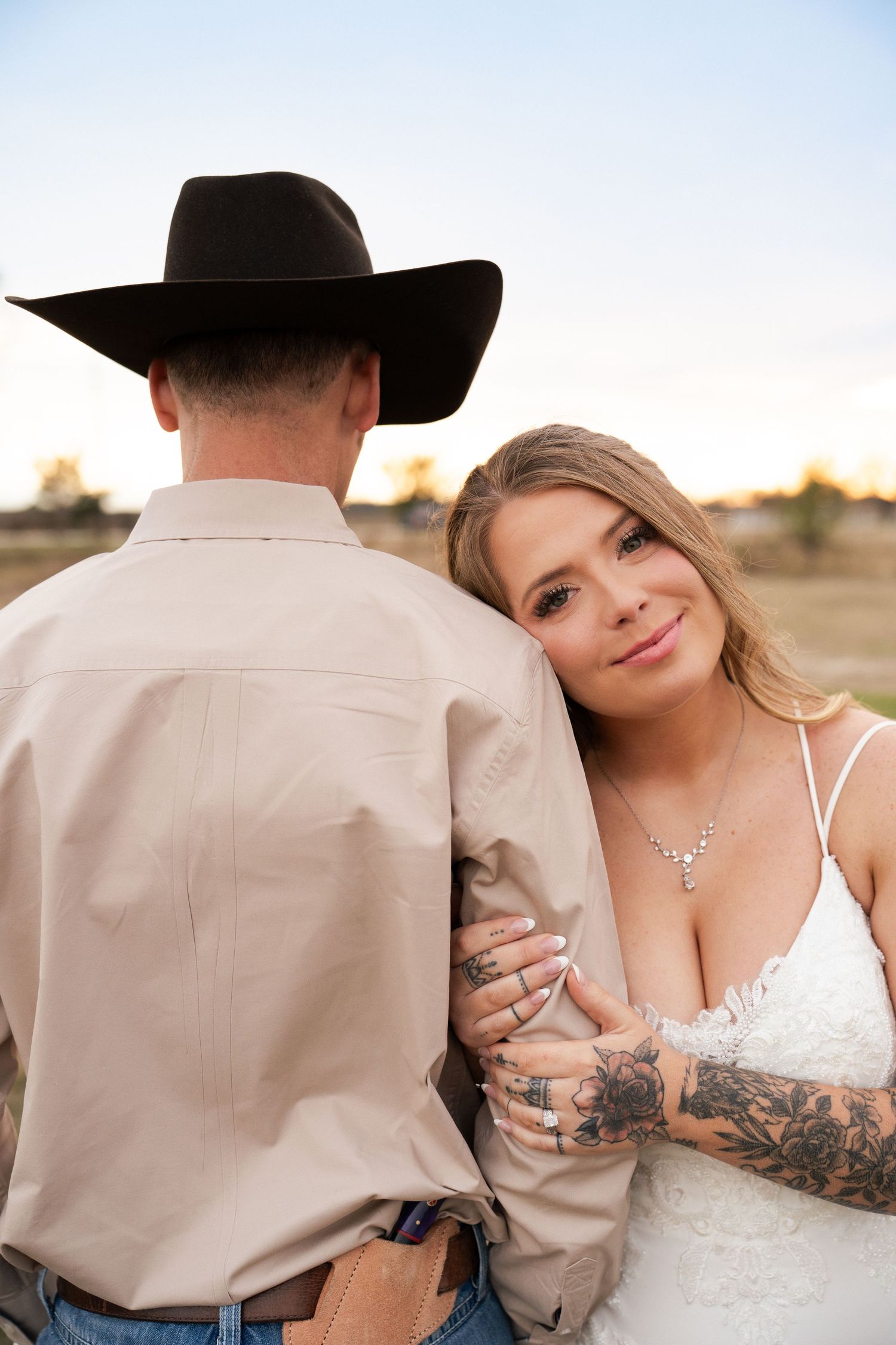 Tattooed bride leans on grooms shoulder as he wears a black cowboy hat at sunset.