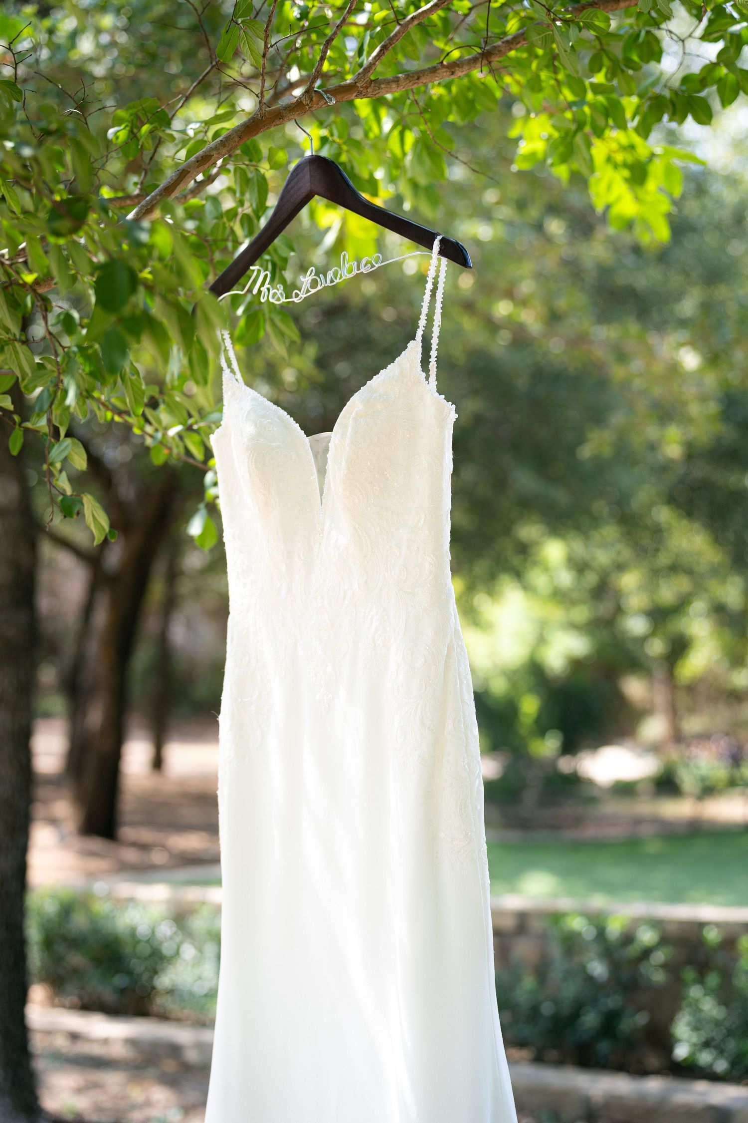 White wedding dress hanging from a tree branch on a custom bridal hanger in a lush outdoor garden setting.