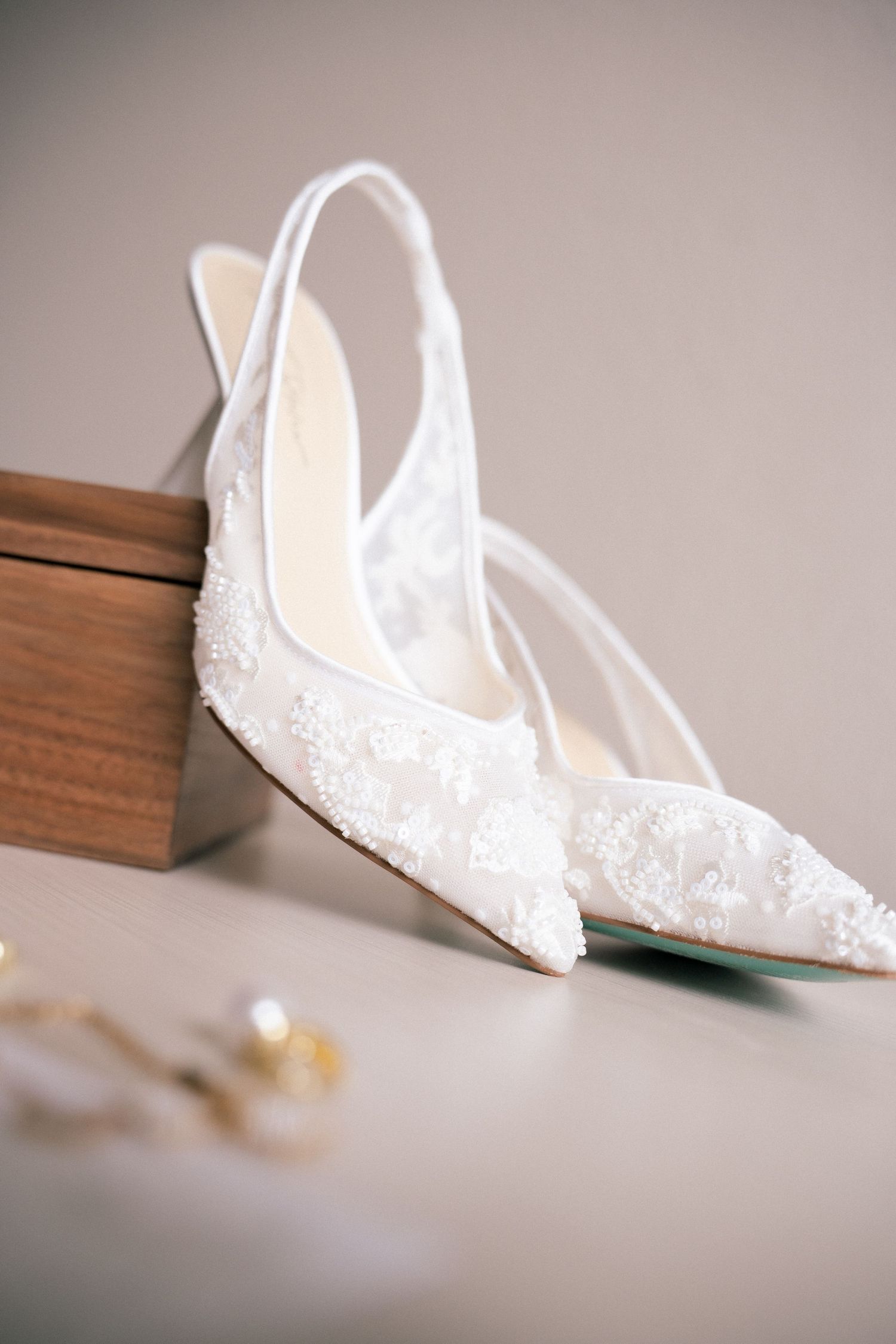 White lace slingback bridal heels with floral embroidery displayed beside a wooden ring box.