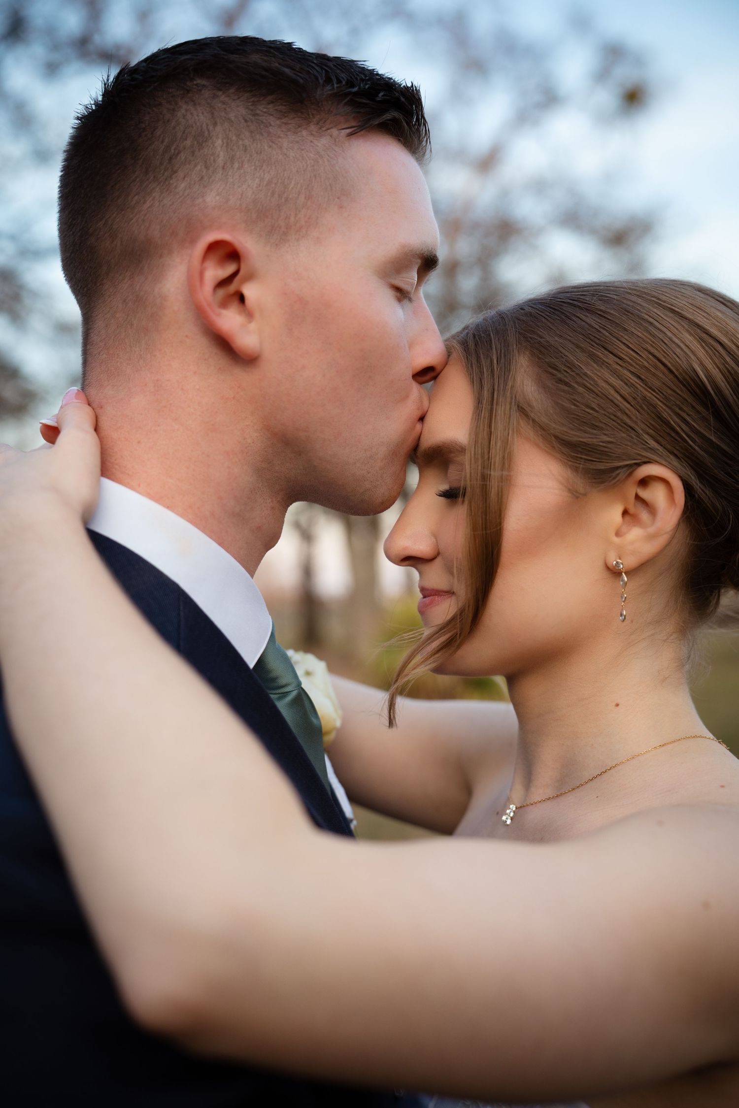 Groom tenderly kisses bride on forehead during an intimate outdoor wedding portrait at dusk.