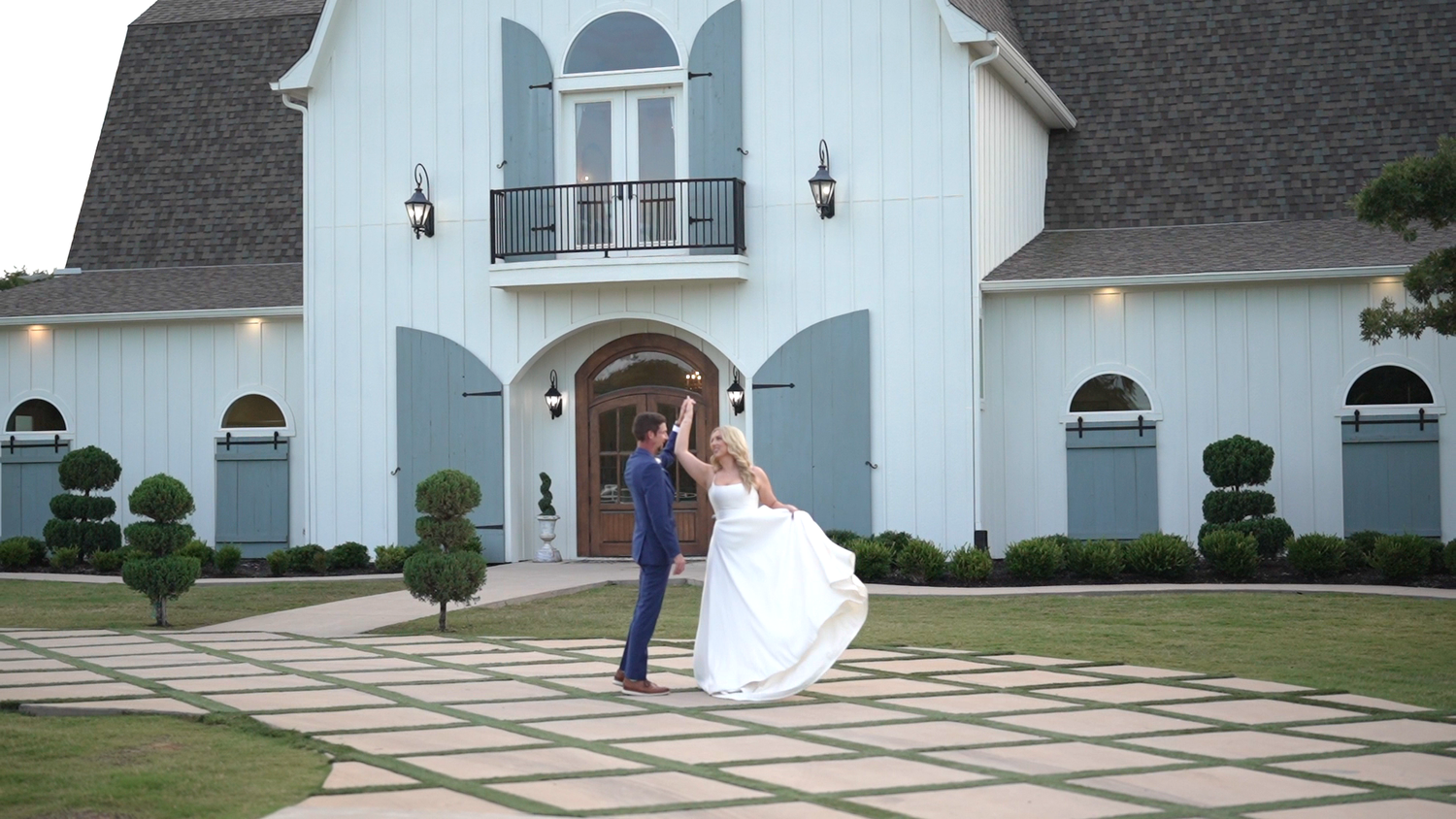 Groom twirls bride in flowing white dress outside a stunning white wedding venue with blue doors.