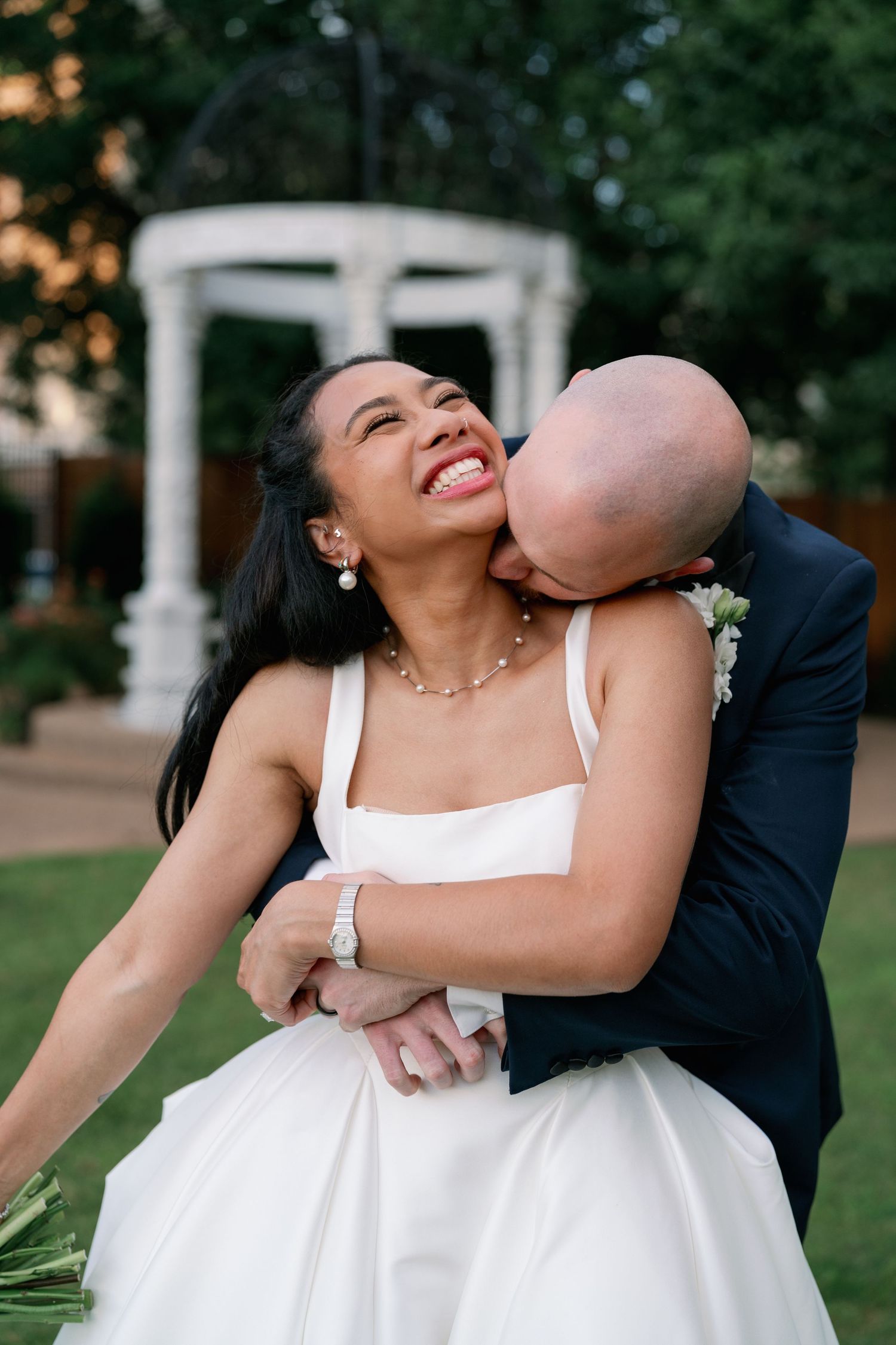 Groom kissing bride on neck while she laughs joyfully at outdoor wedding venue.
