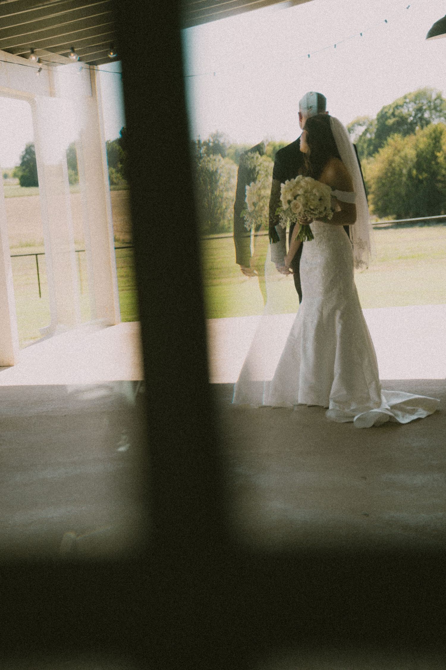Artistic shot of bride in mermaid gown holding bouquet viewed through barn doorway columns.