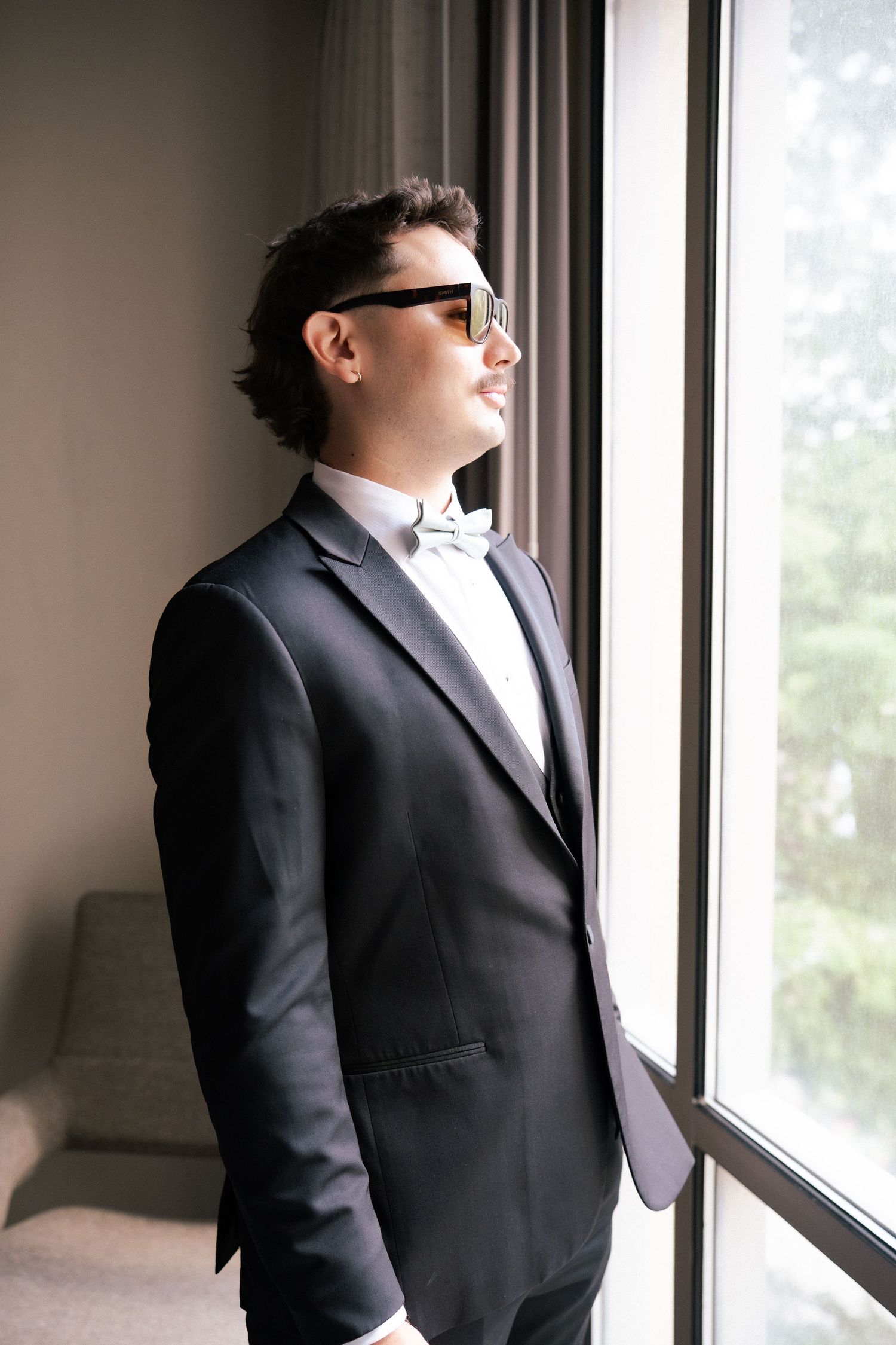 A stylish groom in a black tuxedo and white bow tie wearing sunglasses stands by a large window.