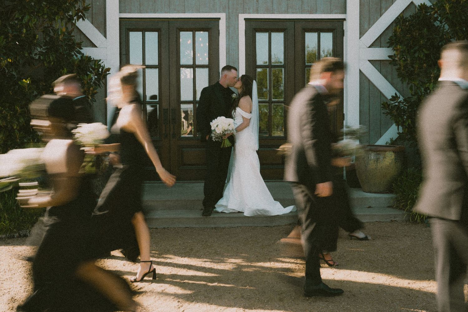 Bride and groom share a kiss outside barn venue as wedding party walks past in motion blur.