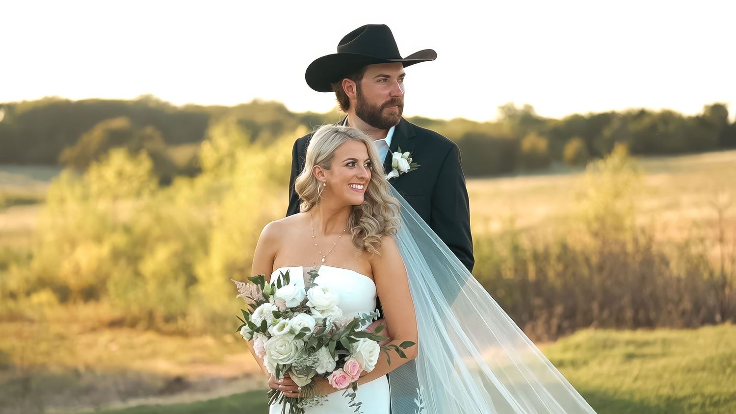 Bride in a strapless gown holds a bouquet while groom in a black cowboy hat embraces her at a golden hour outdoor wedding.