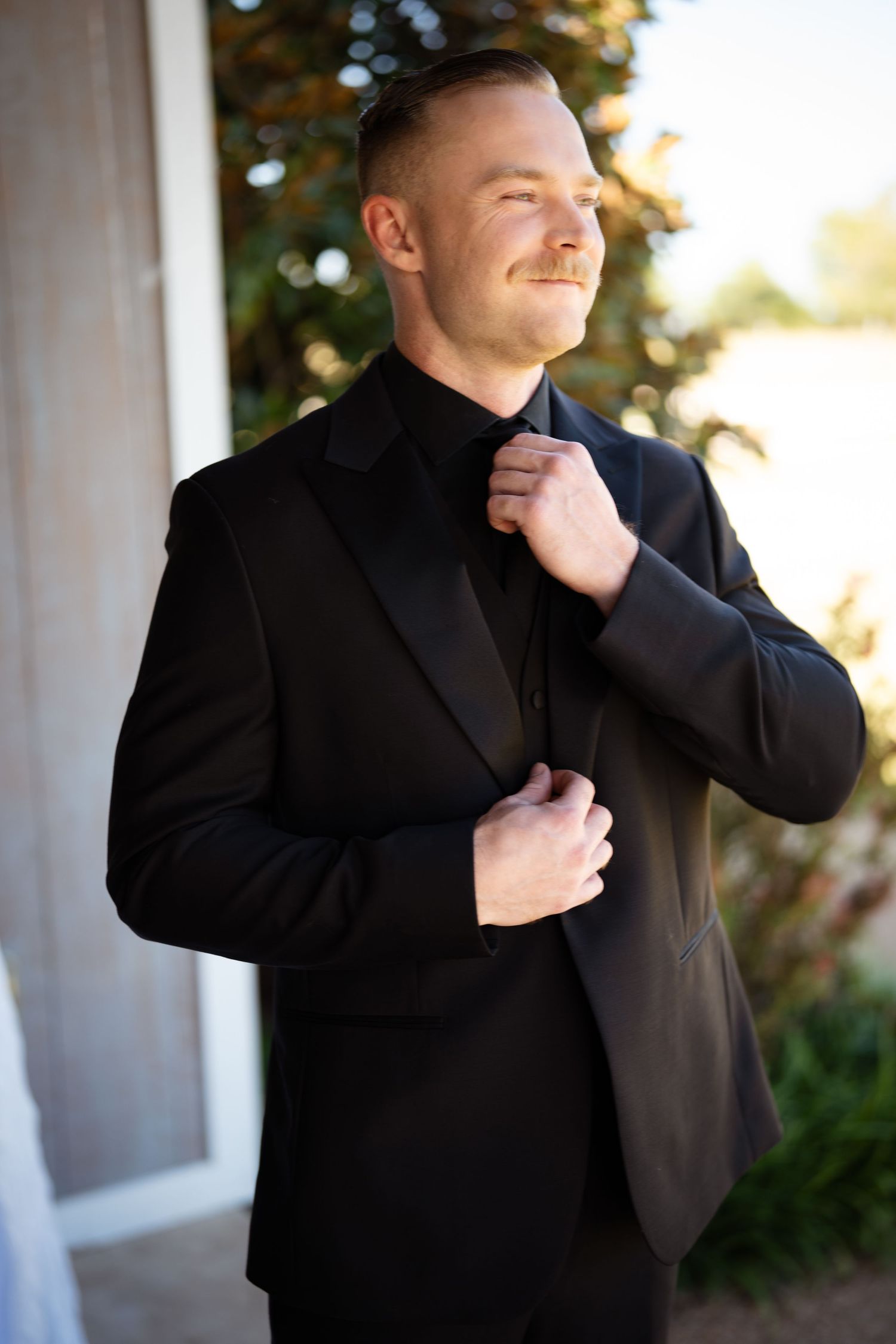 Groom in black tuxedo adjusts bow tie while standing outdoors in natural light.