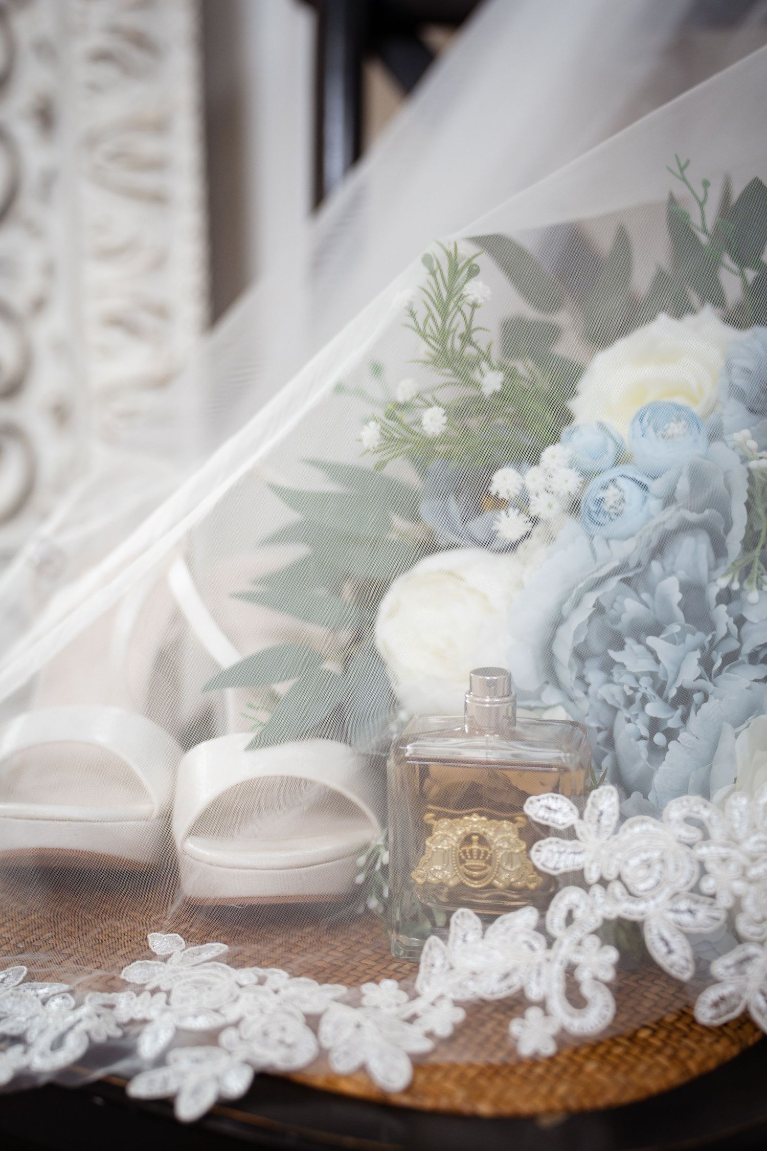 Bridal details flat lay with white heels, blue floral bouquet, perfume bottle, and lace veil.