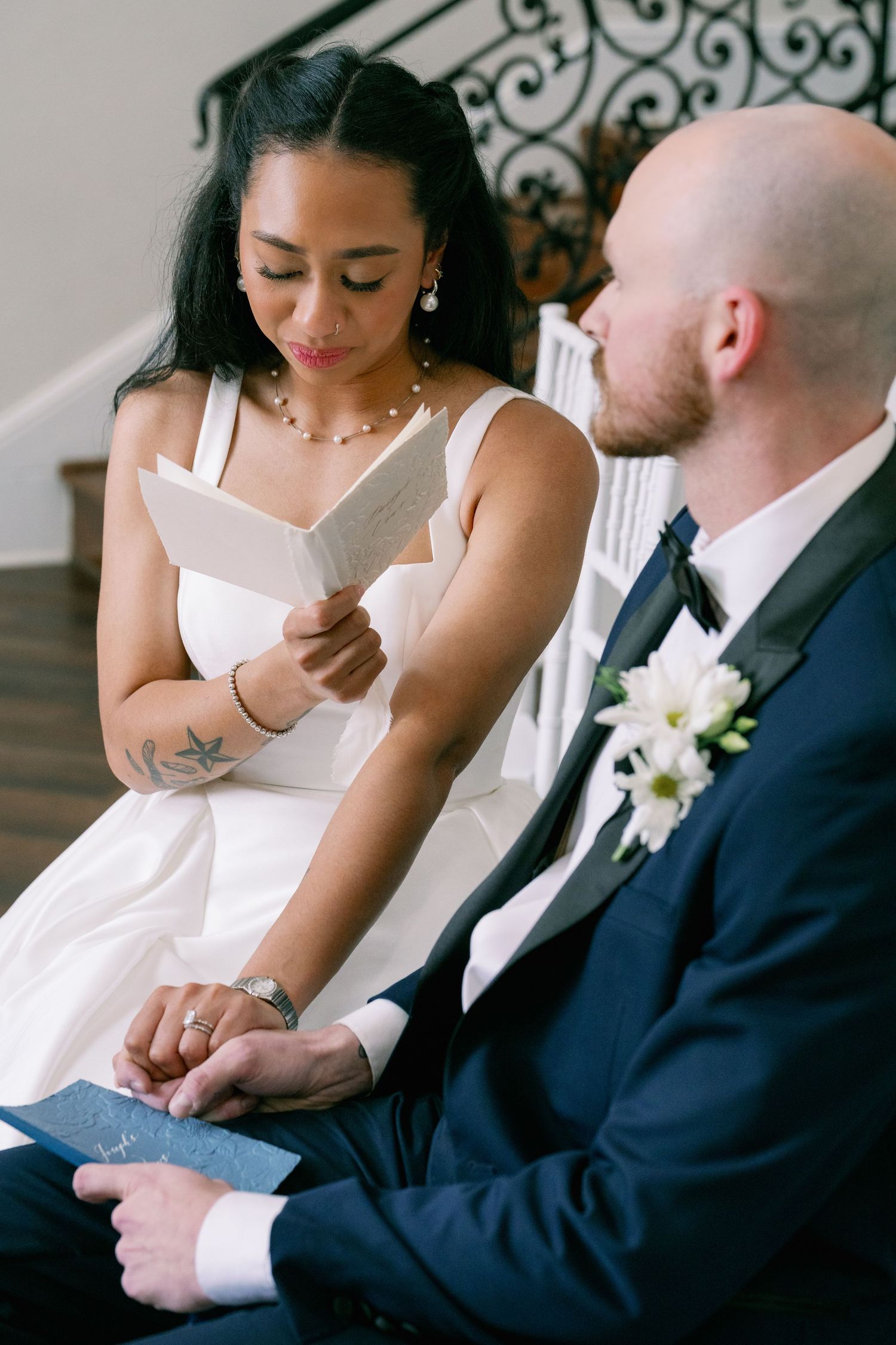 Bride reads heartfelt vows from a card while holding grooms hand during intimate indoor wedding ceremony.