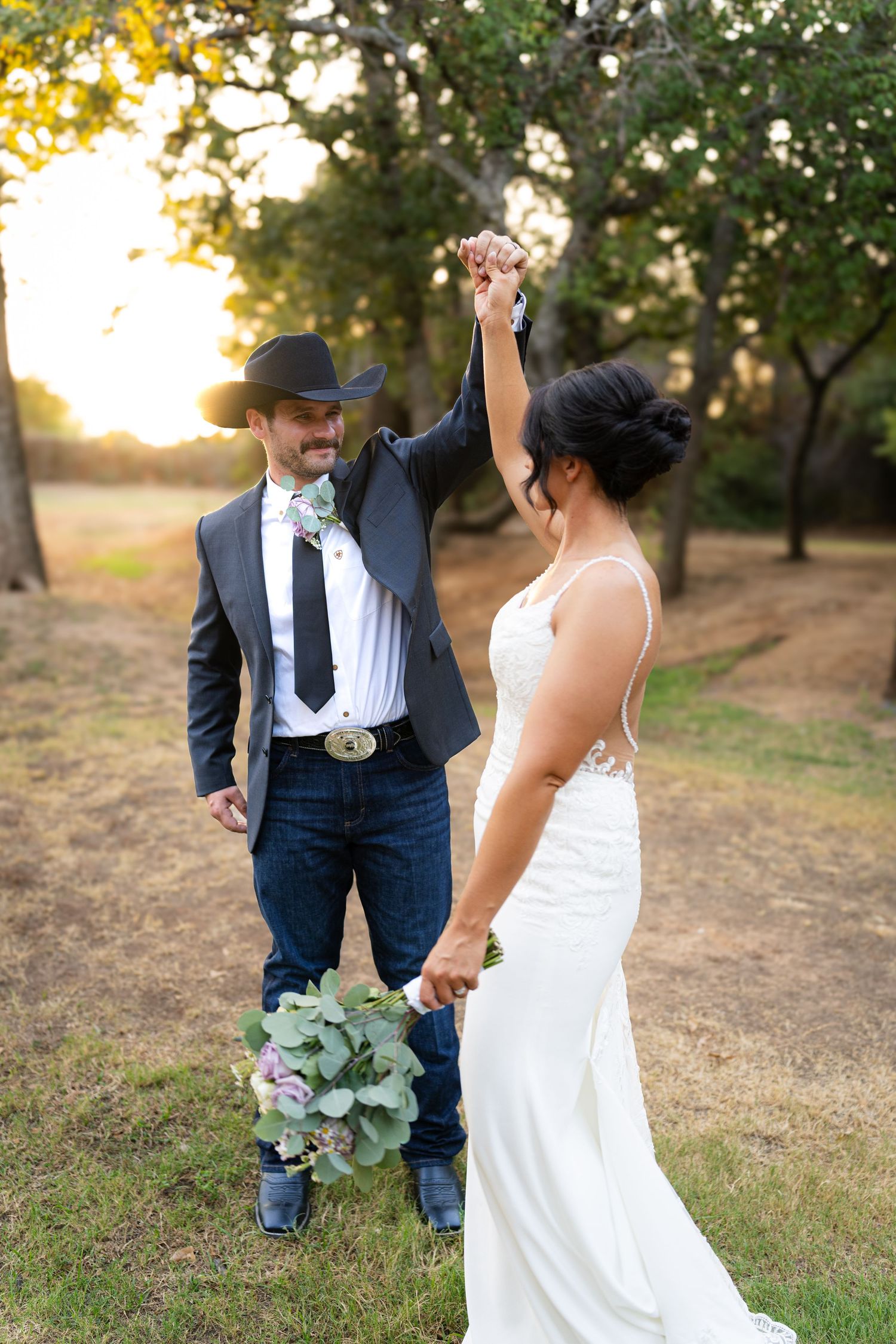 Bride in white lace gown and groom in cowboy hat and jeans dancing outdoors at golden hour wedding.