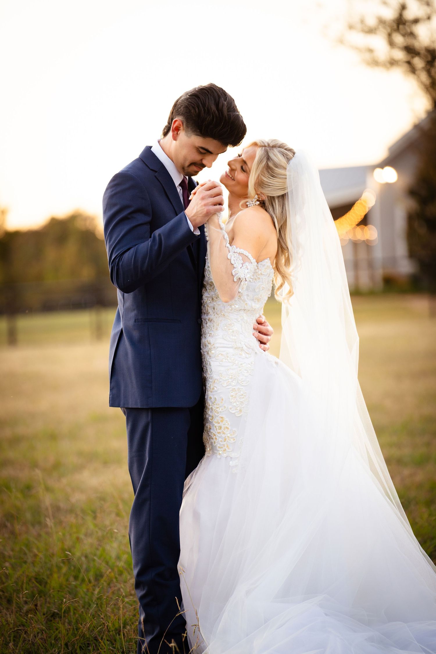 Bride and groom share a romantic golden hour moment outdoors, groom kissing bride's hand in a field.