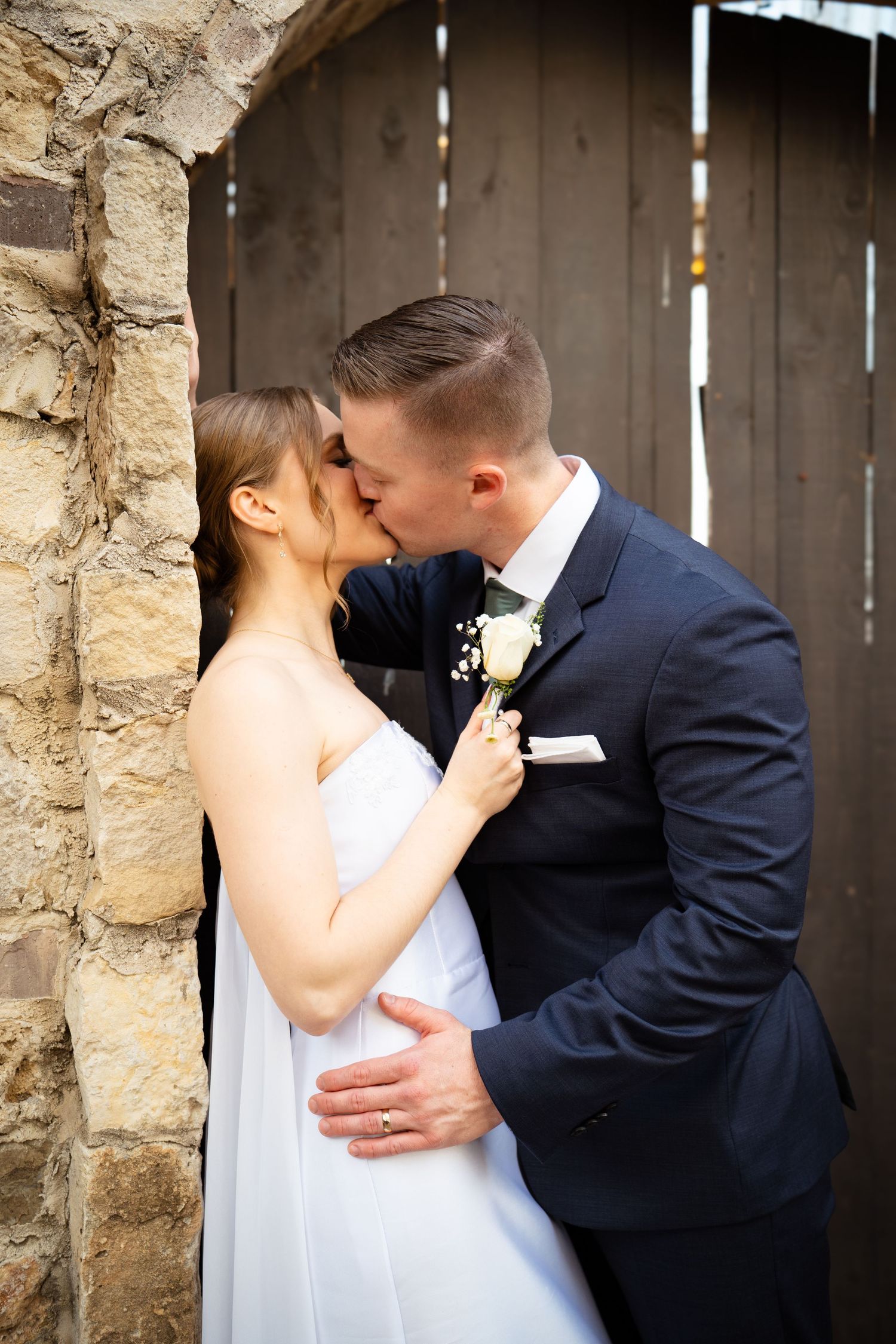 Bride and groom share a passionate kiss against a rustic stone wall and wooden door backdrop.