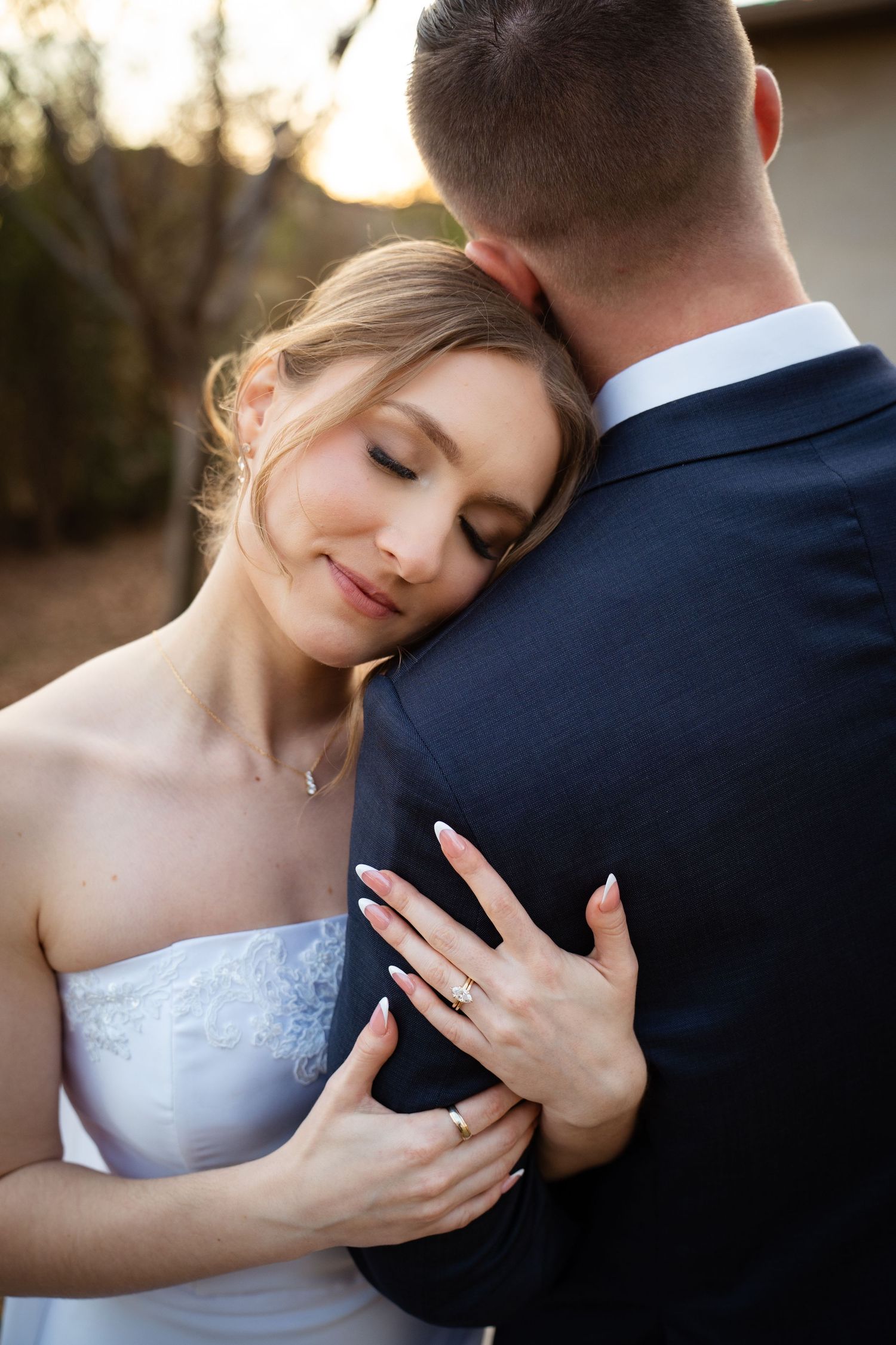 Bride rests her head peacefully on groom's chest, showcasing her wedding ring during sunset portraits.