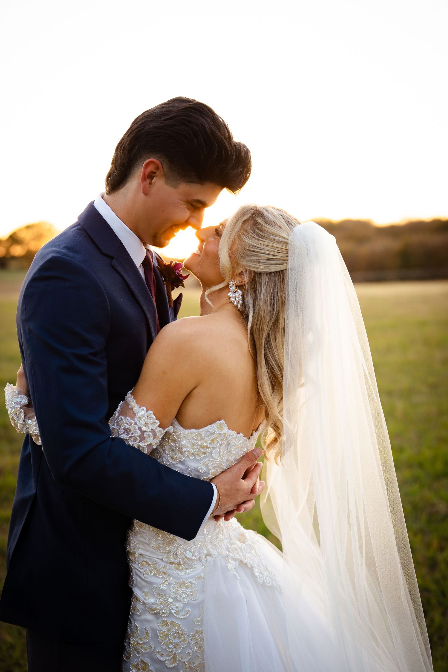 Bride and groom share an intimate embrace at sunset in an open field, foreheads nearly touching.