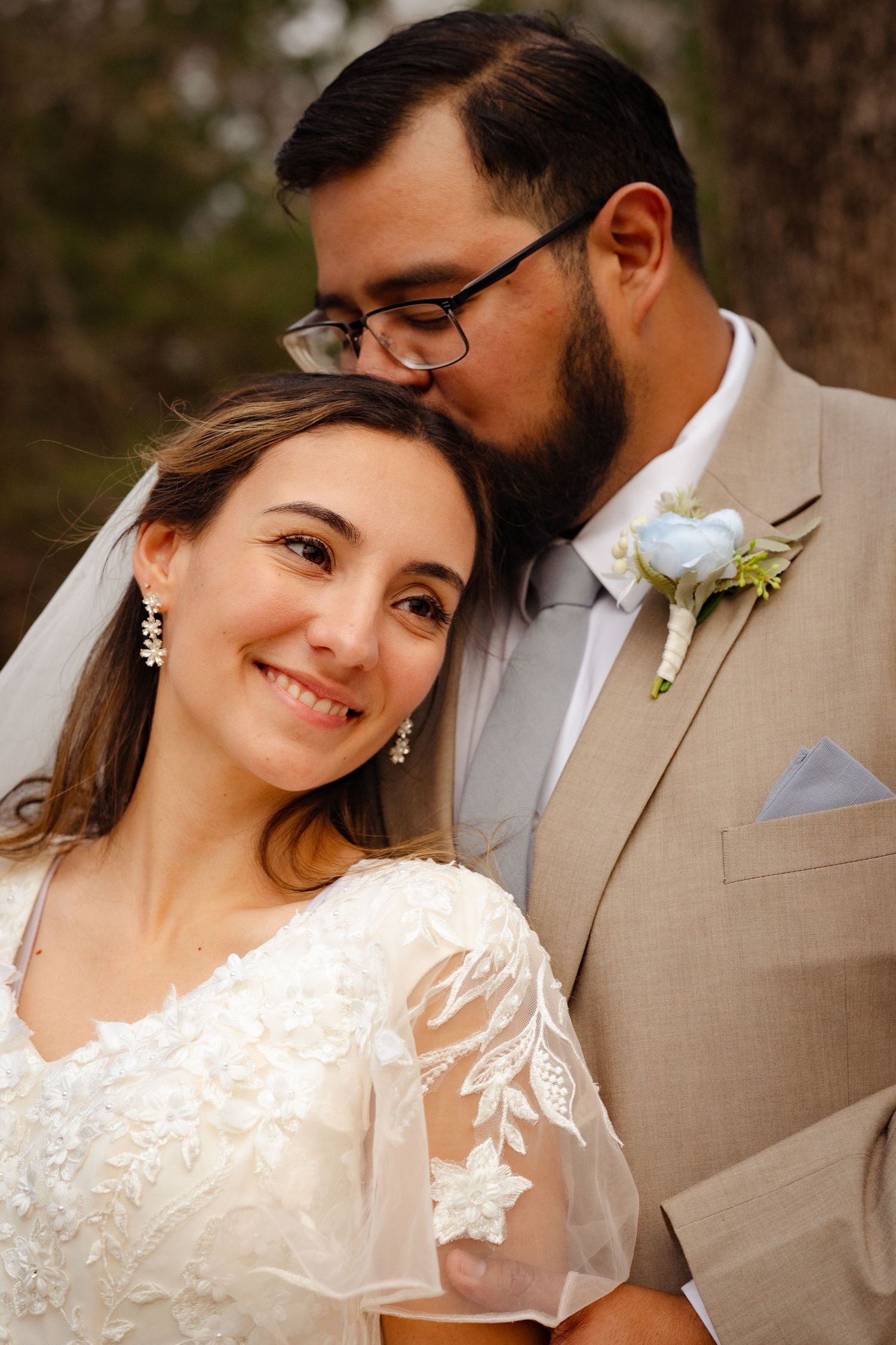 Groom in tan suit kissing bride on forehead as she smiles warmly at camera outdoors.