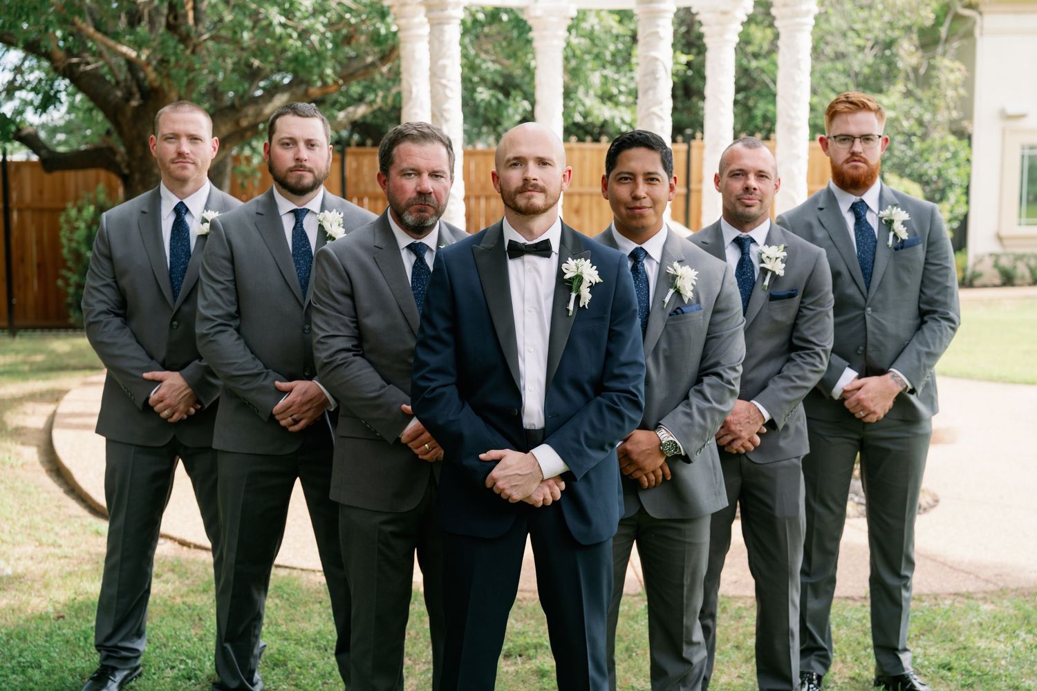 Groom in navy tuxedo stands confidently with six groomsmen in gray suits at outdoor wedding venue.
