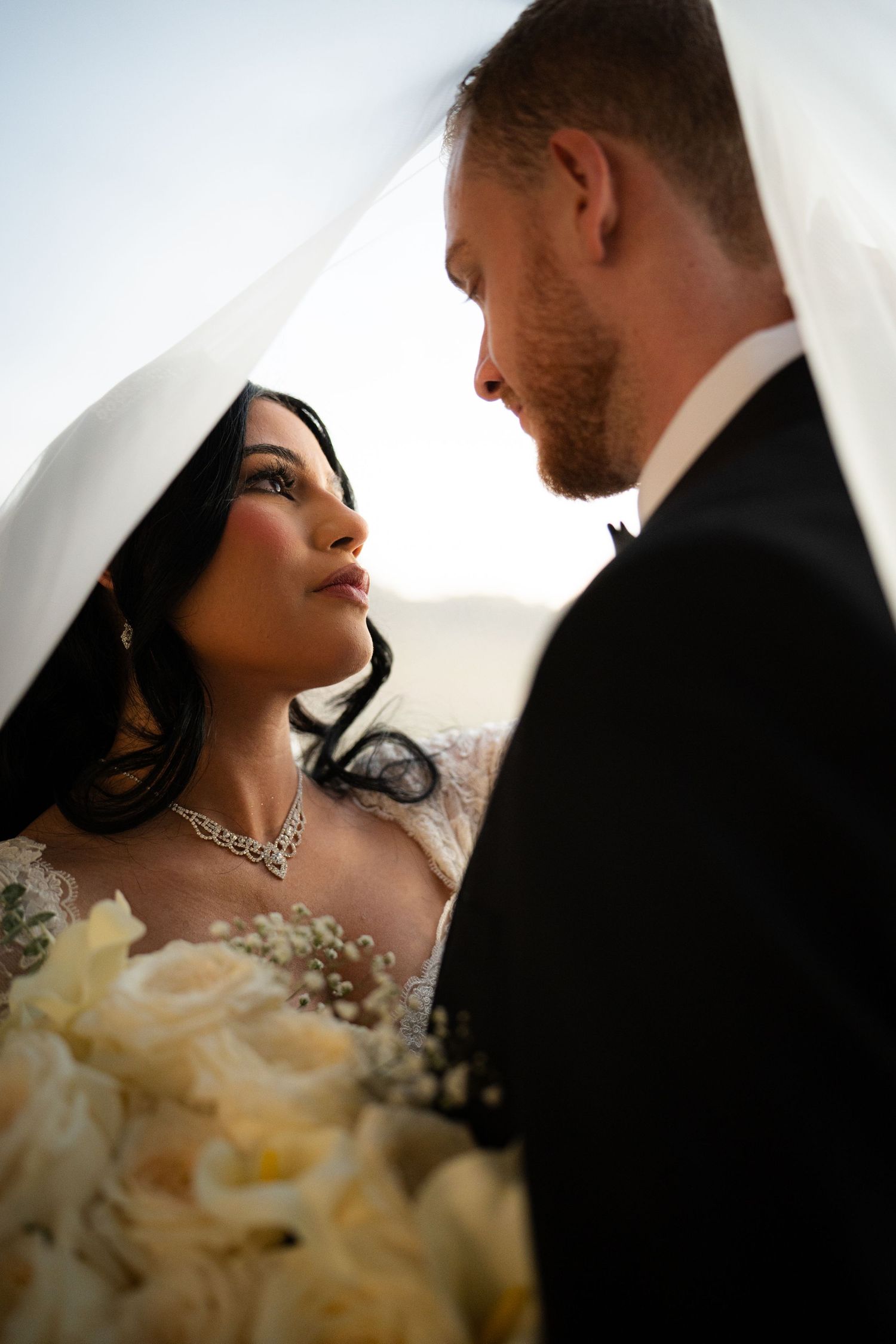 Bride and groom sharing an intimate moment under a veil with white rose bouquet.