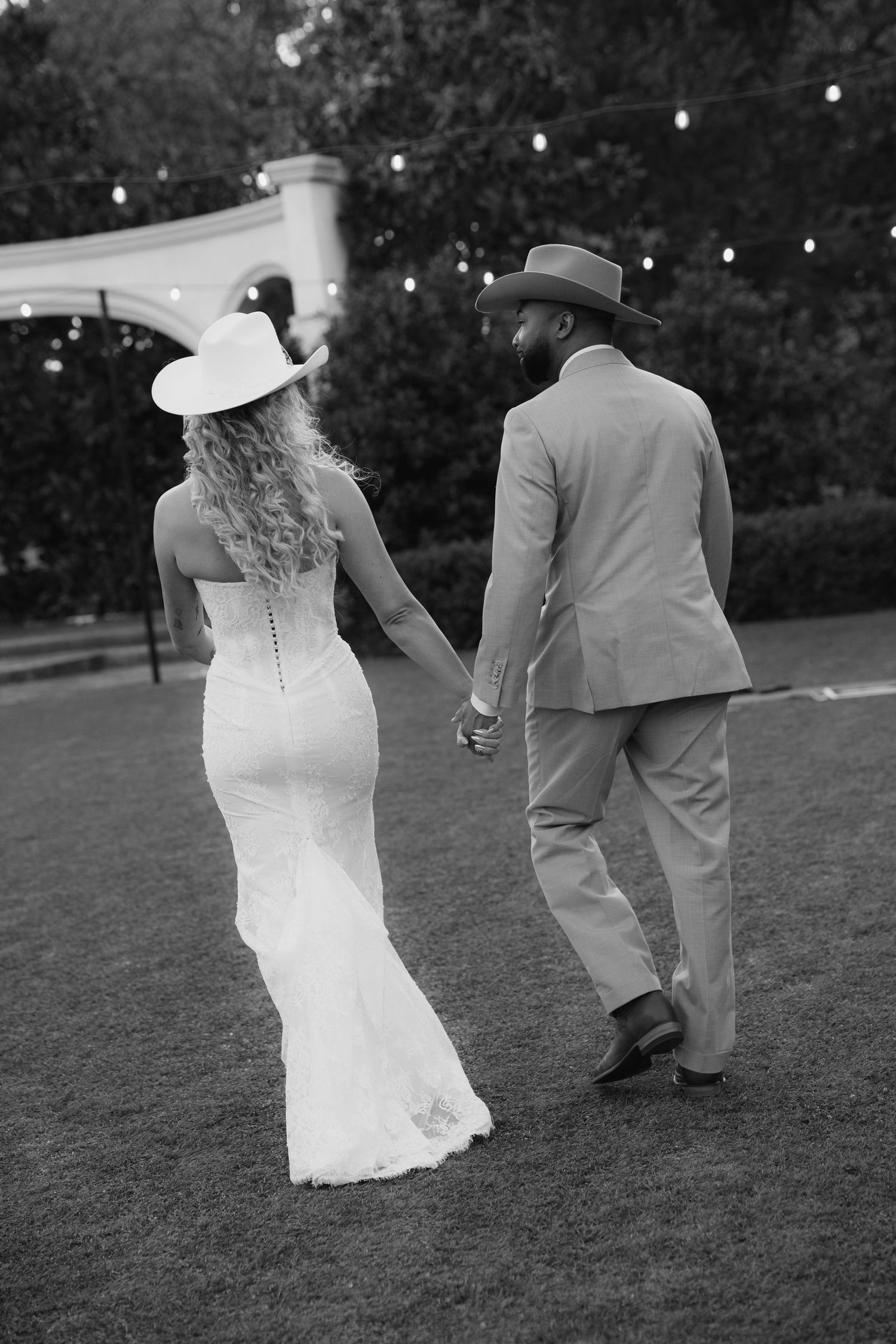 Black and white photo of bride and groom in cowboy hats walking hand in hand across lawn under string lights.