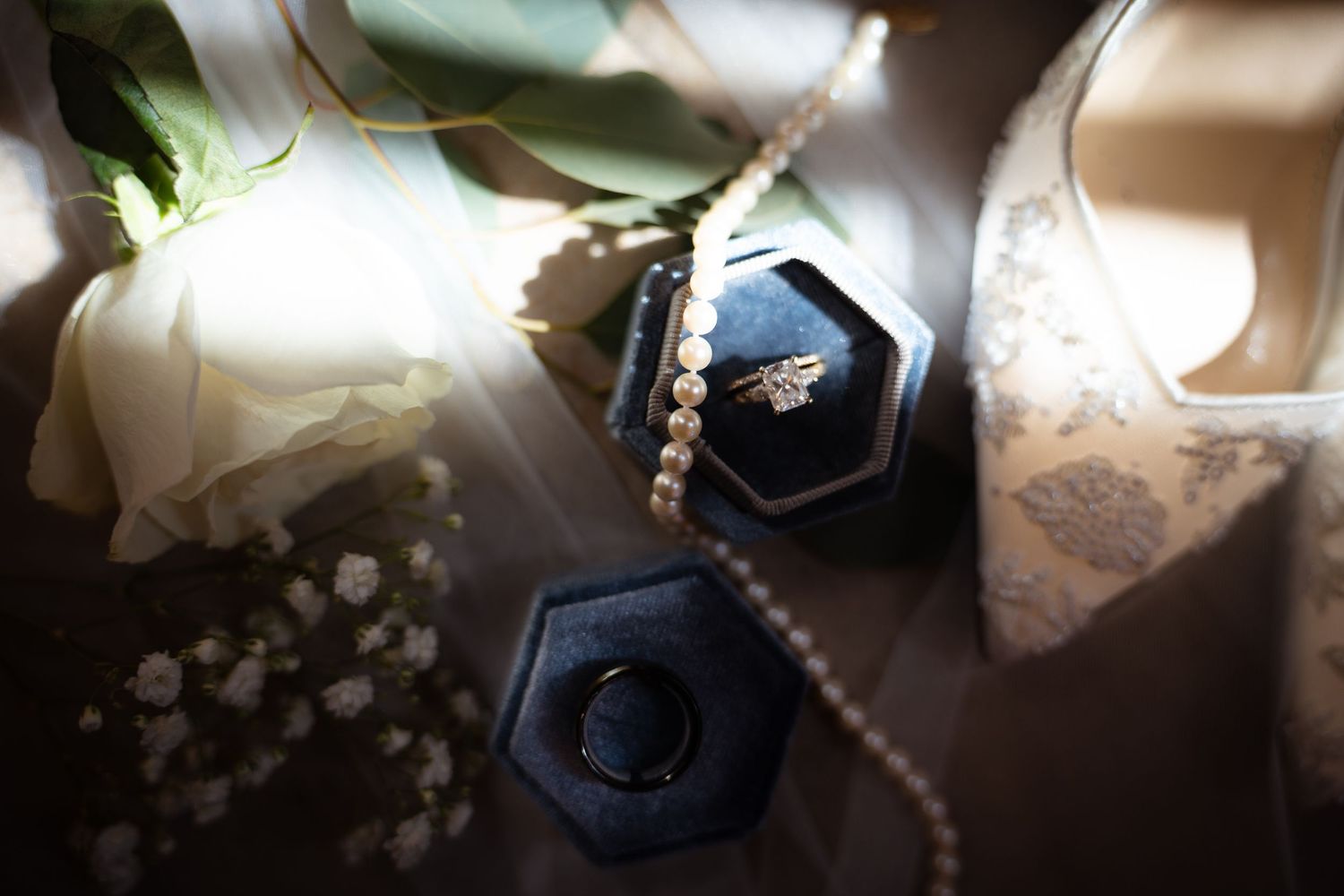 Elegant wedding flat lay featuring diamond rings in velvet box, pearl necklace, and white roses.