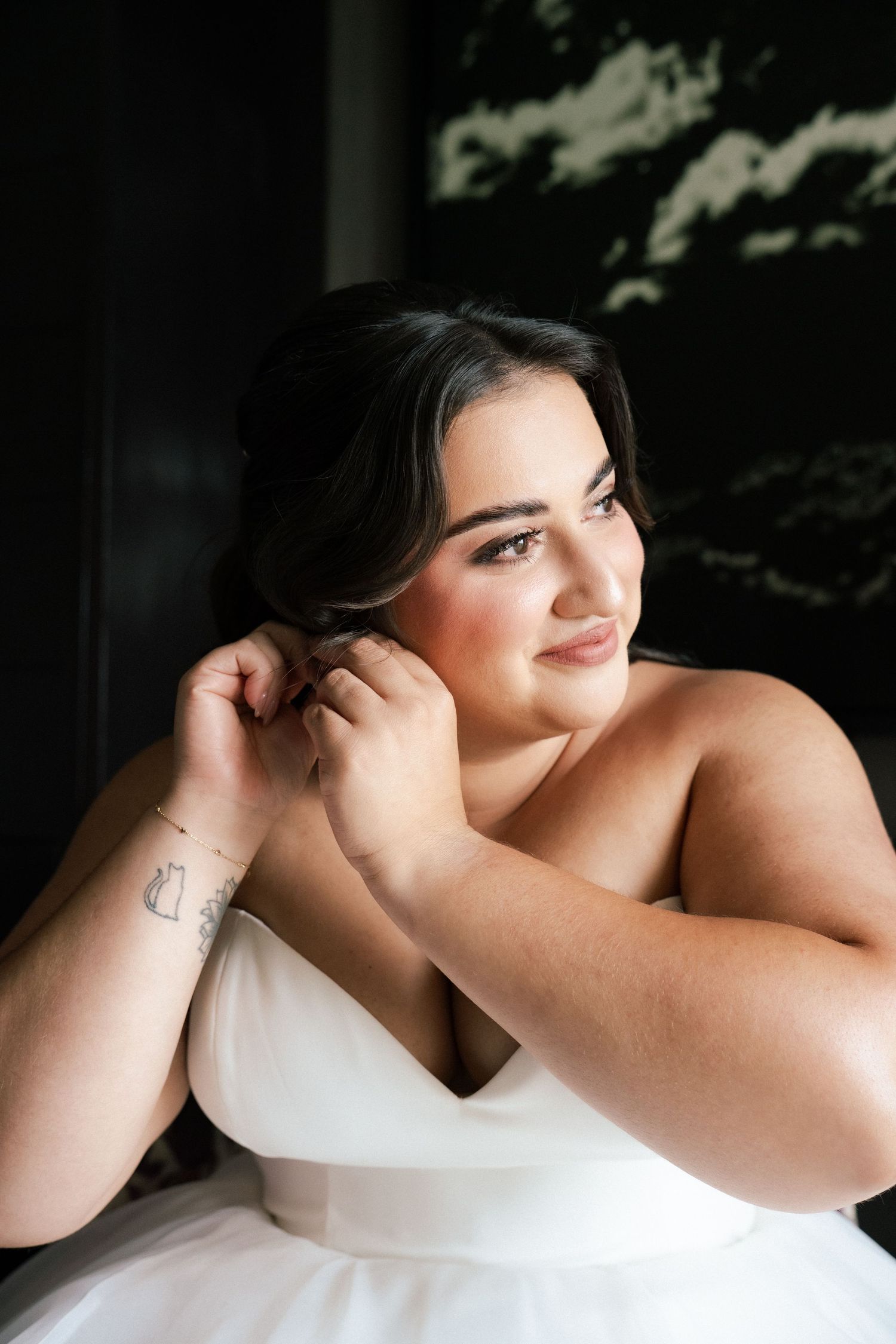 A bride in a white strapless gown adjusts her earring while sitting near a dark window on her wedding day.
