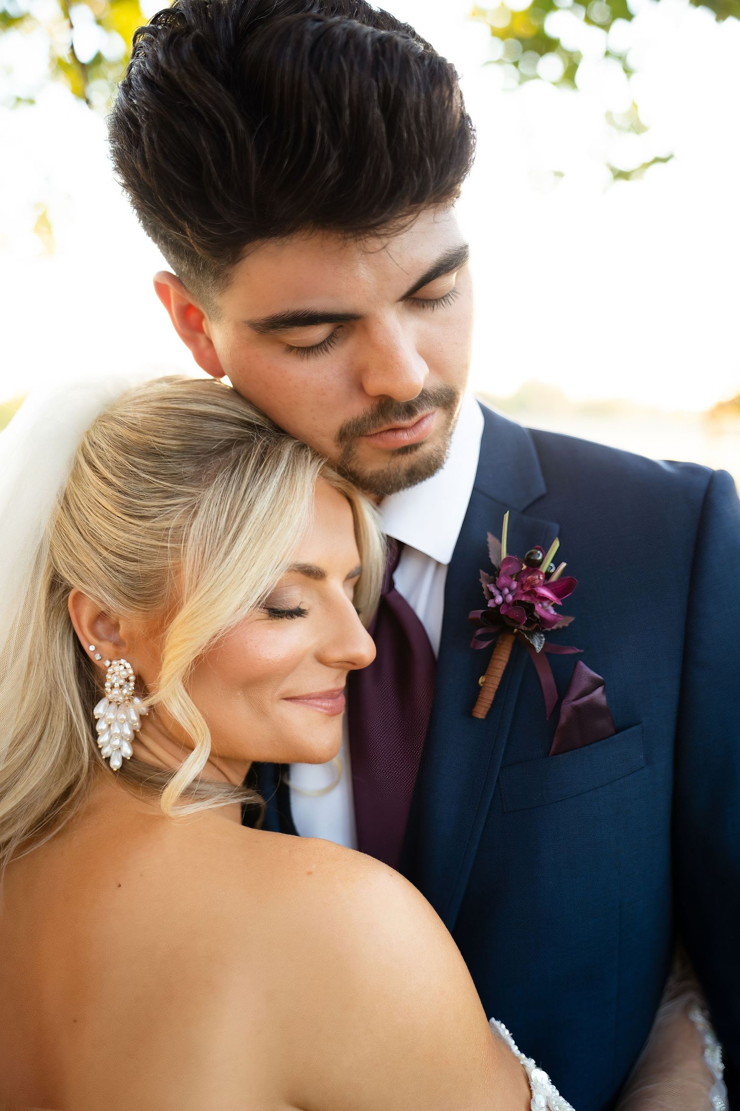 Groom in navy suit with burgundy boutonniere holds bride close in a tender outdoor portrait.