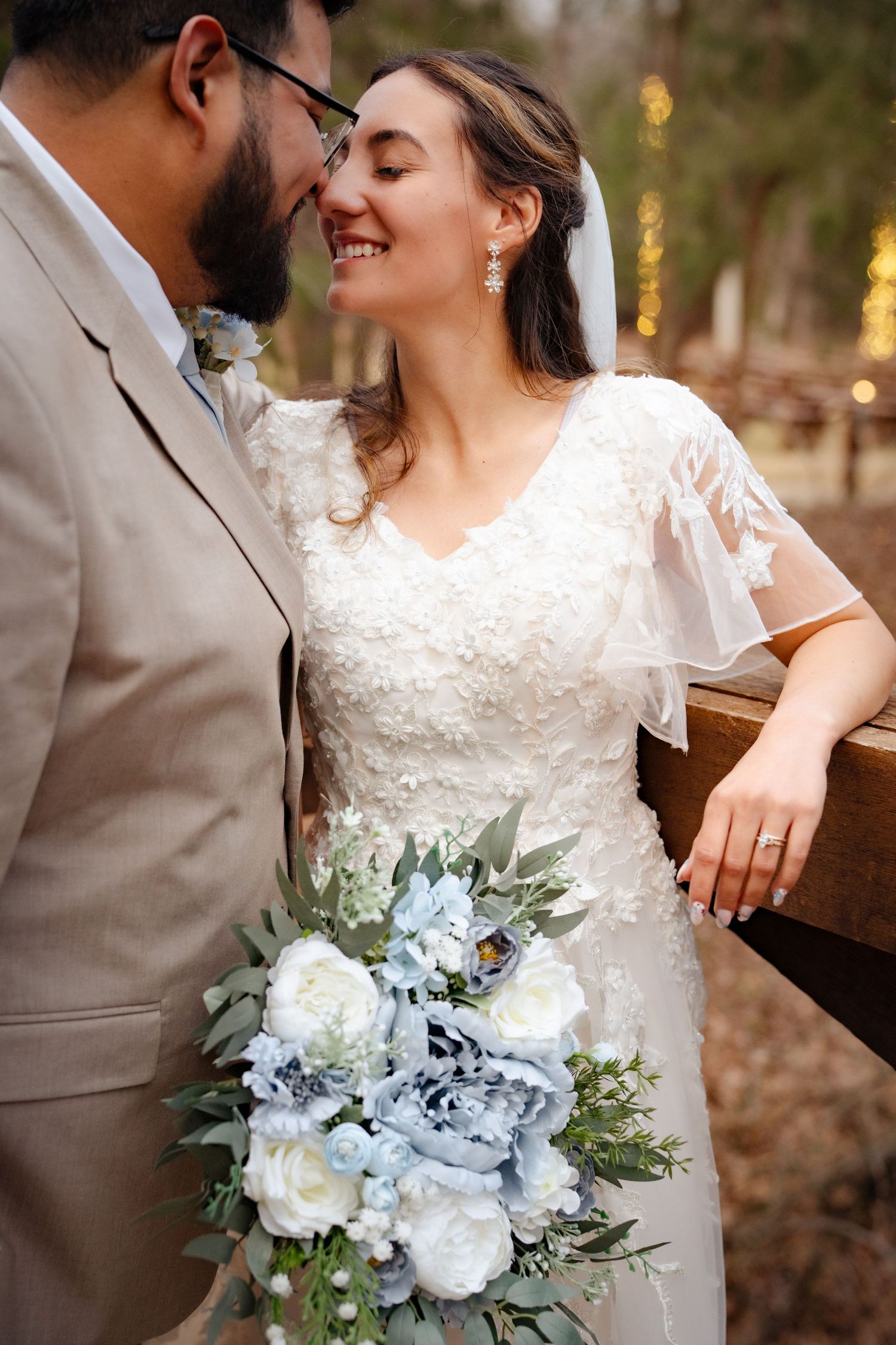 Bride holding blue and white bouquet leans close to bearded groom in tan suit outdoors.
