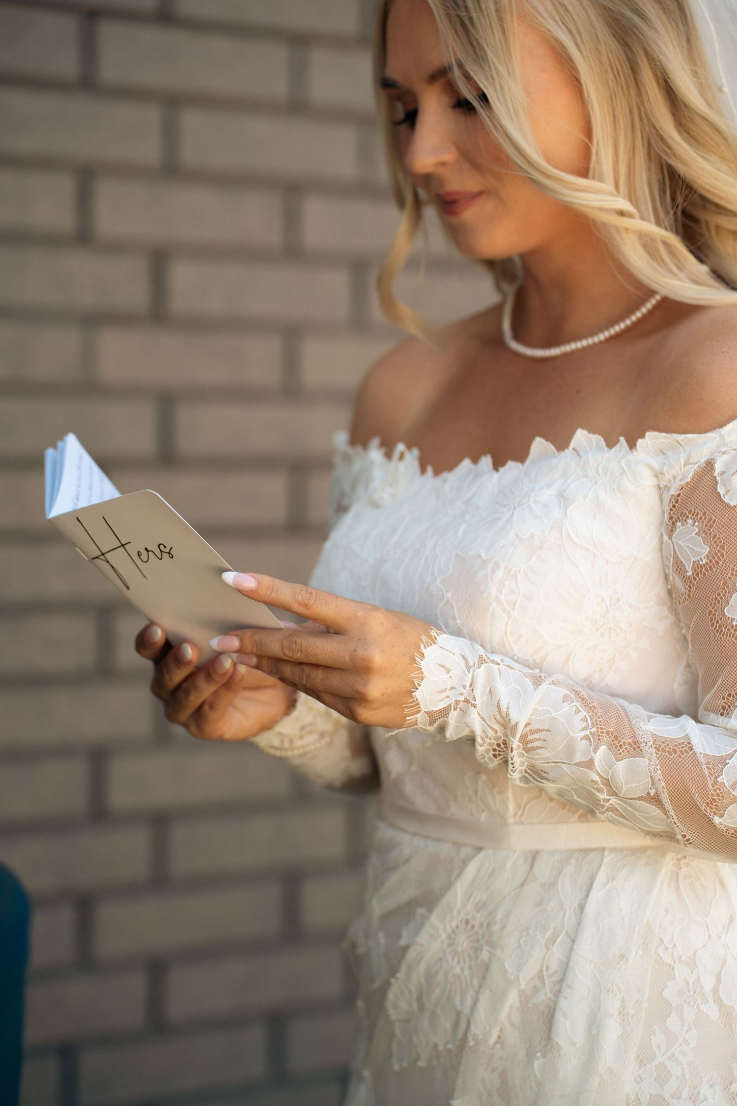 Blonde bride in off-shoulder lace gown reads handwritten vow card while wearing pearl necklace.