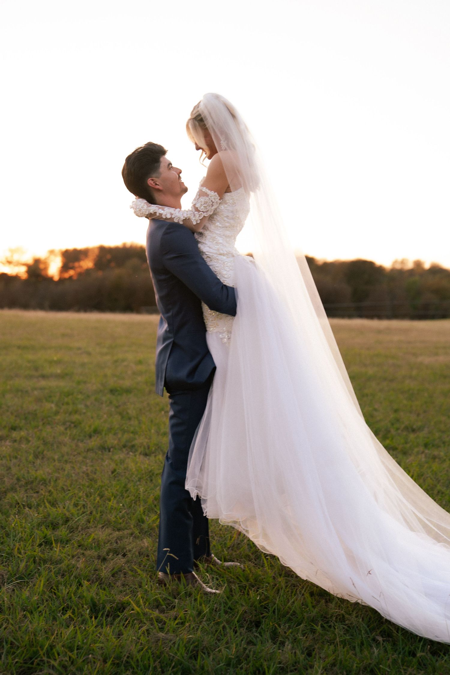 Groom lifts bride in white tulle gown at golden sunset in an open green field.