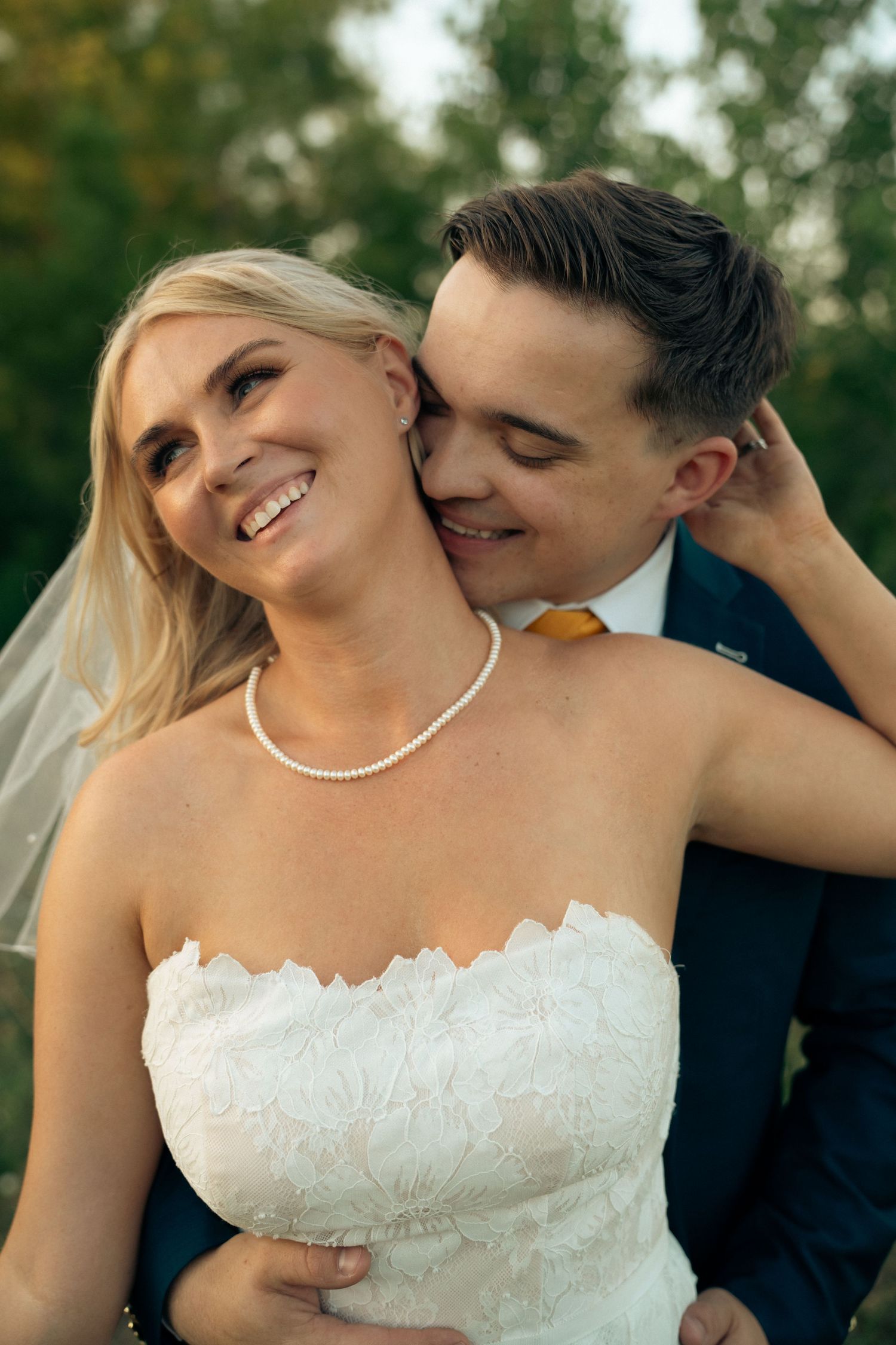 Laughing blonde bride in strapless lace gown with pearl necklace embraced by groom in blue suit.