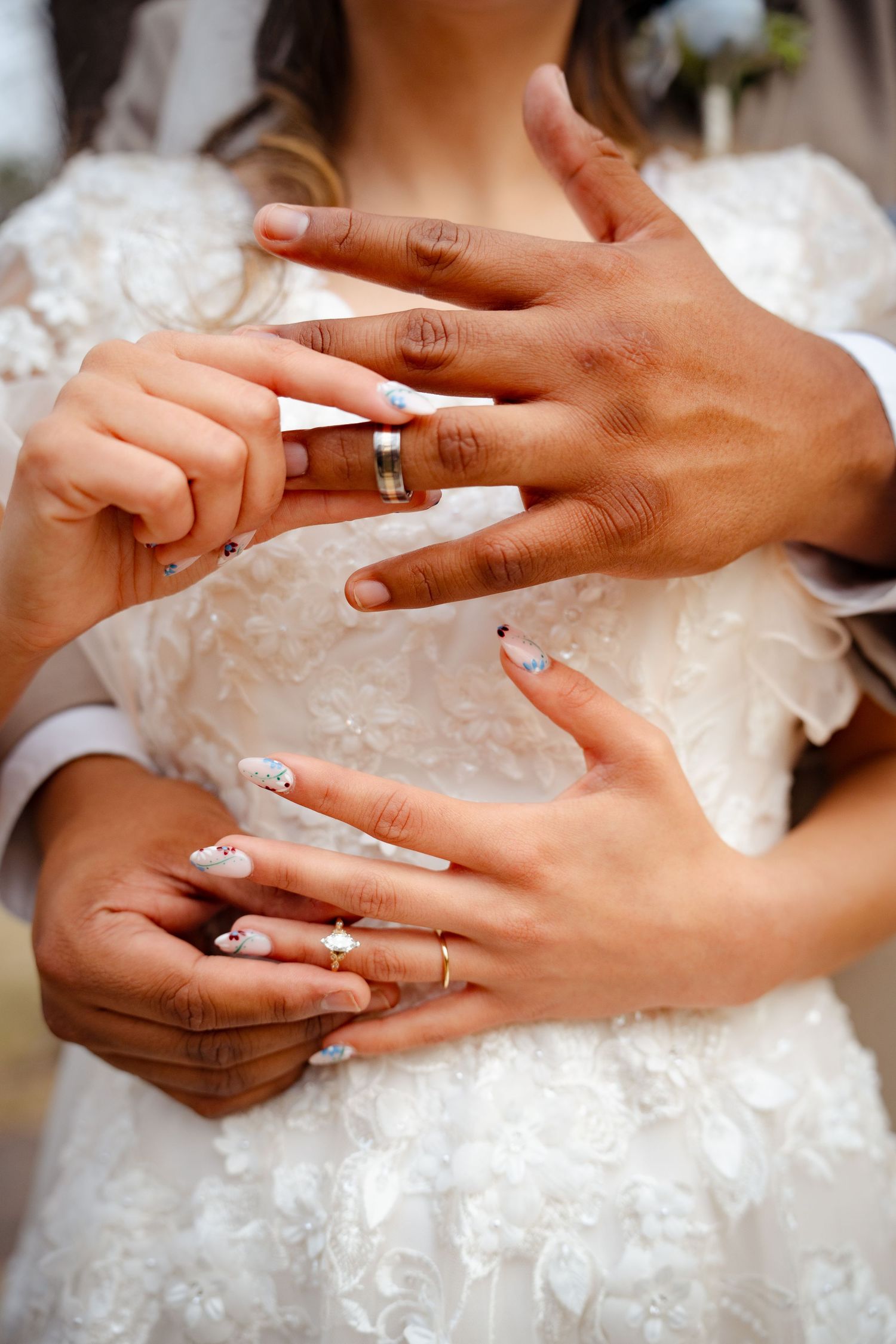 Close-up of interracial newlyweds' hands showing wedding bands and floral nail art over a white lace bridal gown.