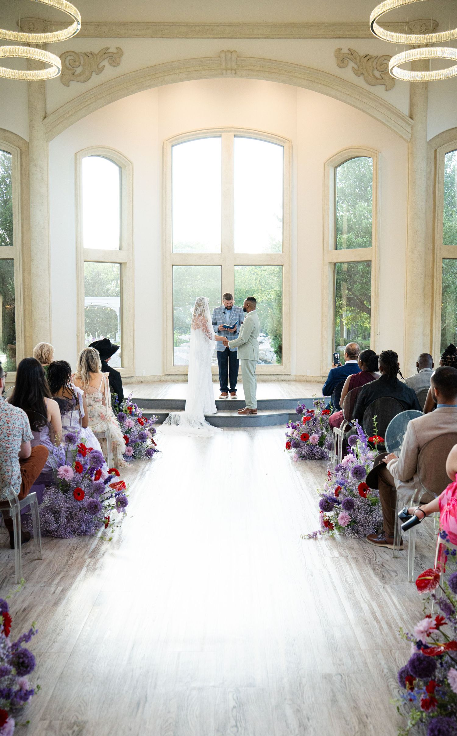 Elegant indoor wedding ceremony with purple floral aisle, tall arched windows, and guests seated in rows.