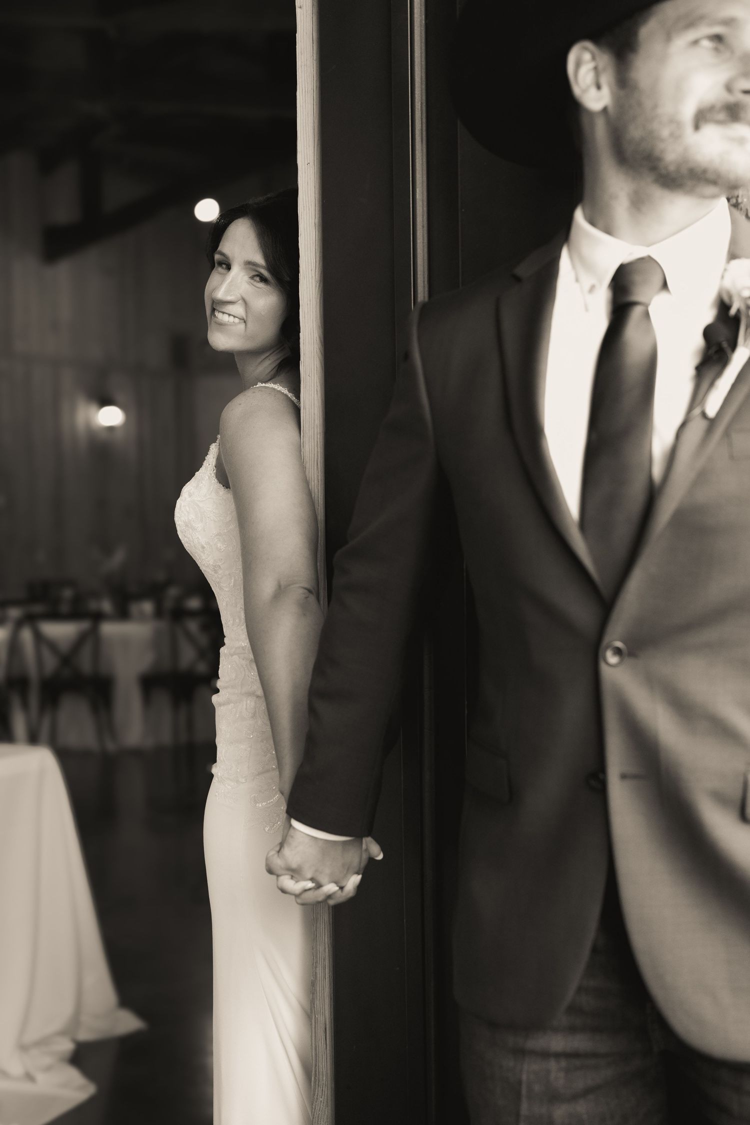 Black and white photo of a bride and groom holding hands around a door pillar at their wedding reception.