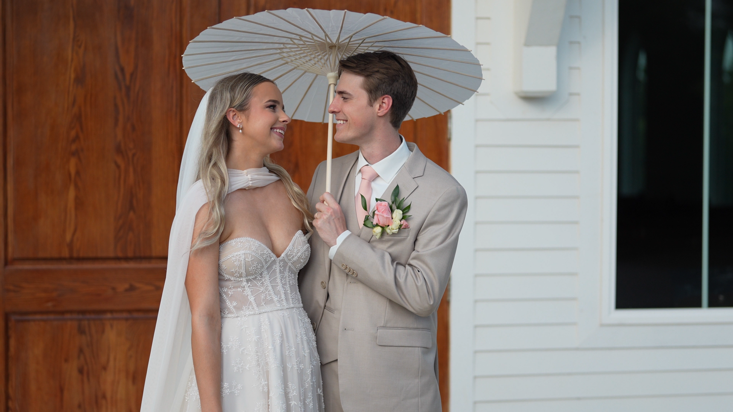 Smiling bride and groom share a moment under a white parasol on their wedding day.