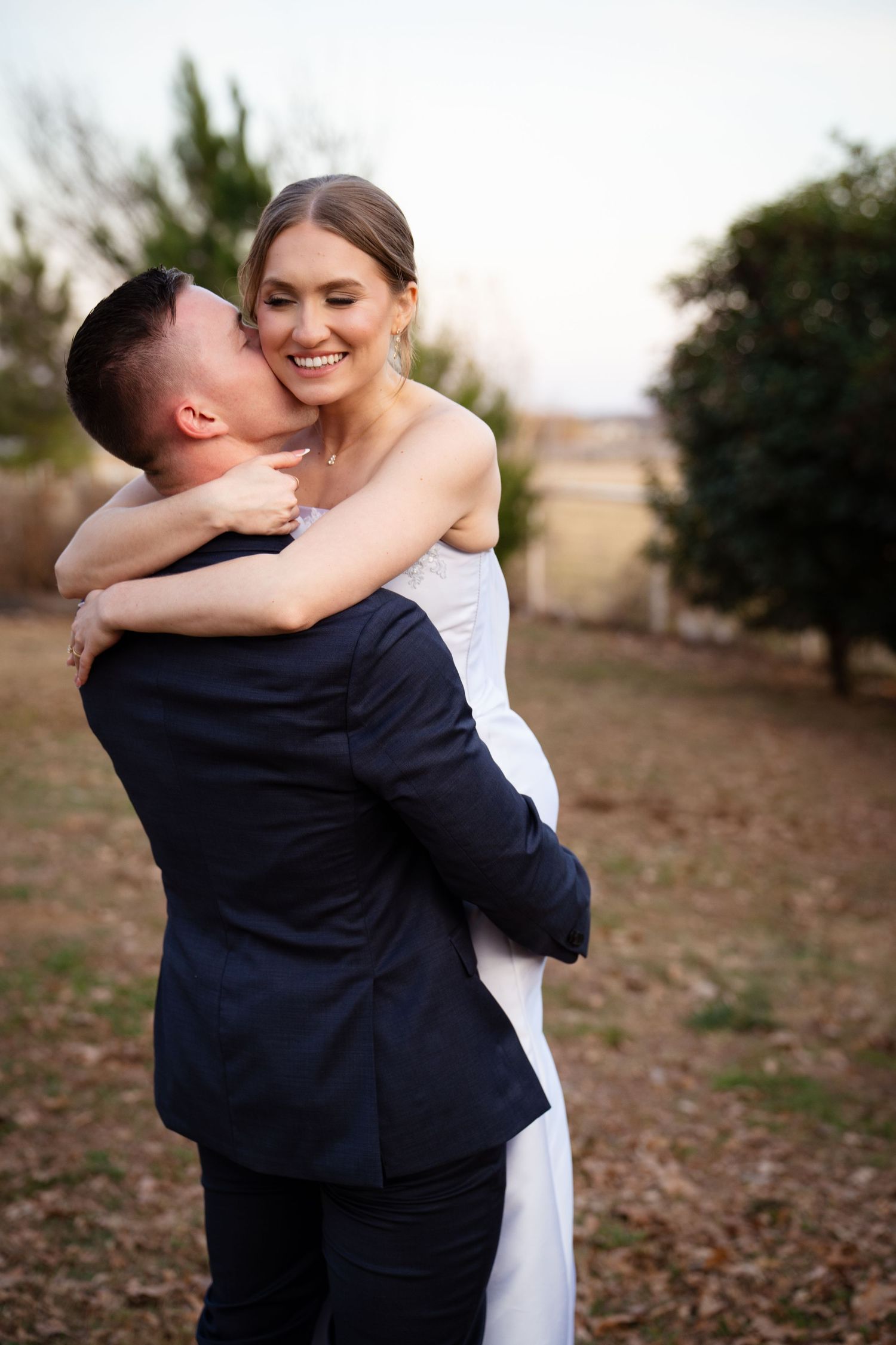 Joyful bride laughs as groom kisses her cheek while holding her close during outdoor wedding photos.