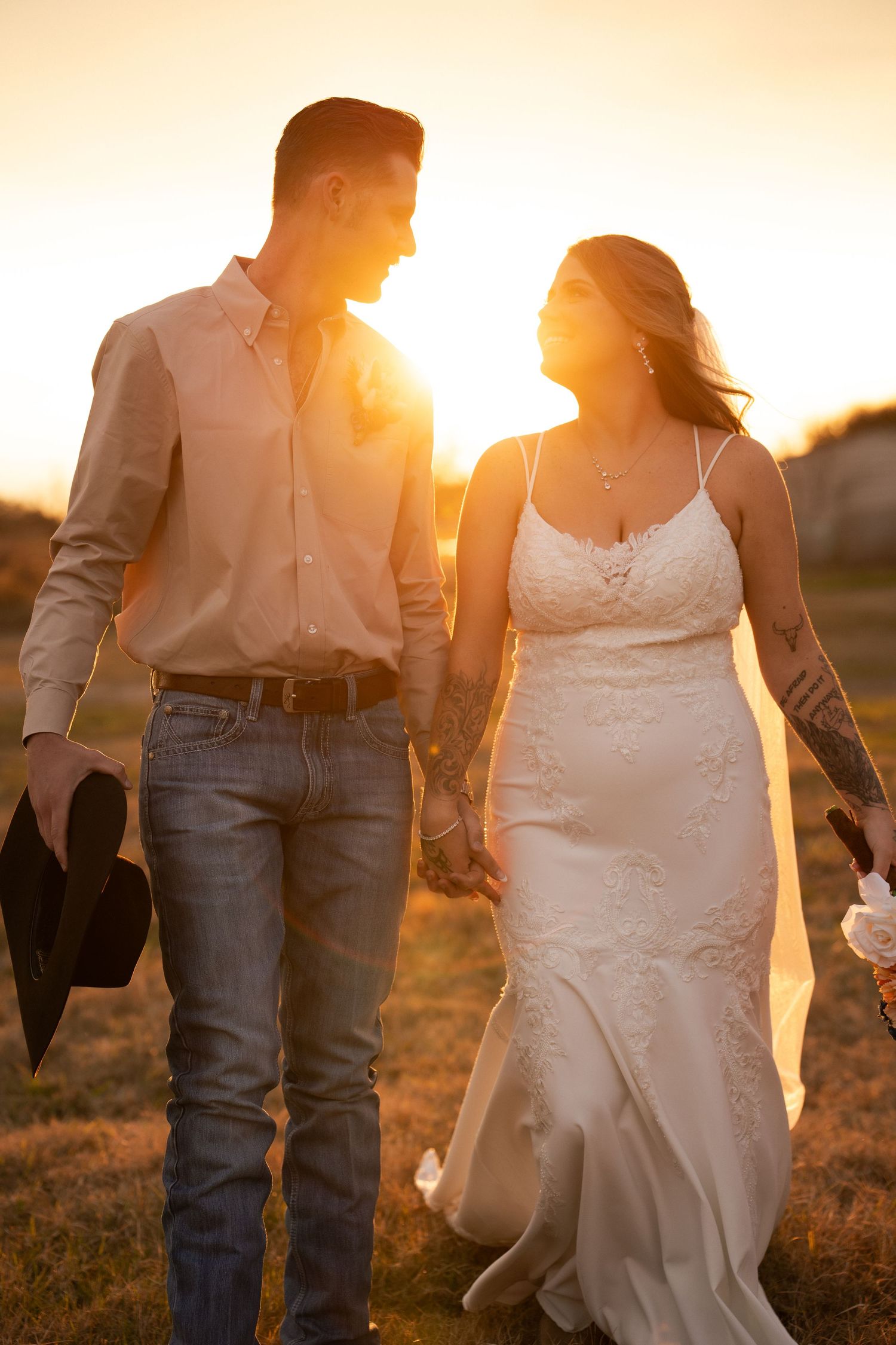 Bride and groom walk hand in hand through golden field at stunning sunset holding hats.