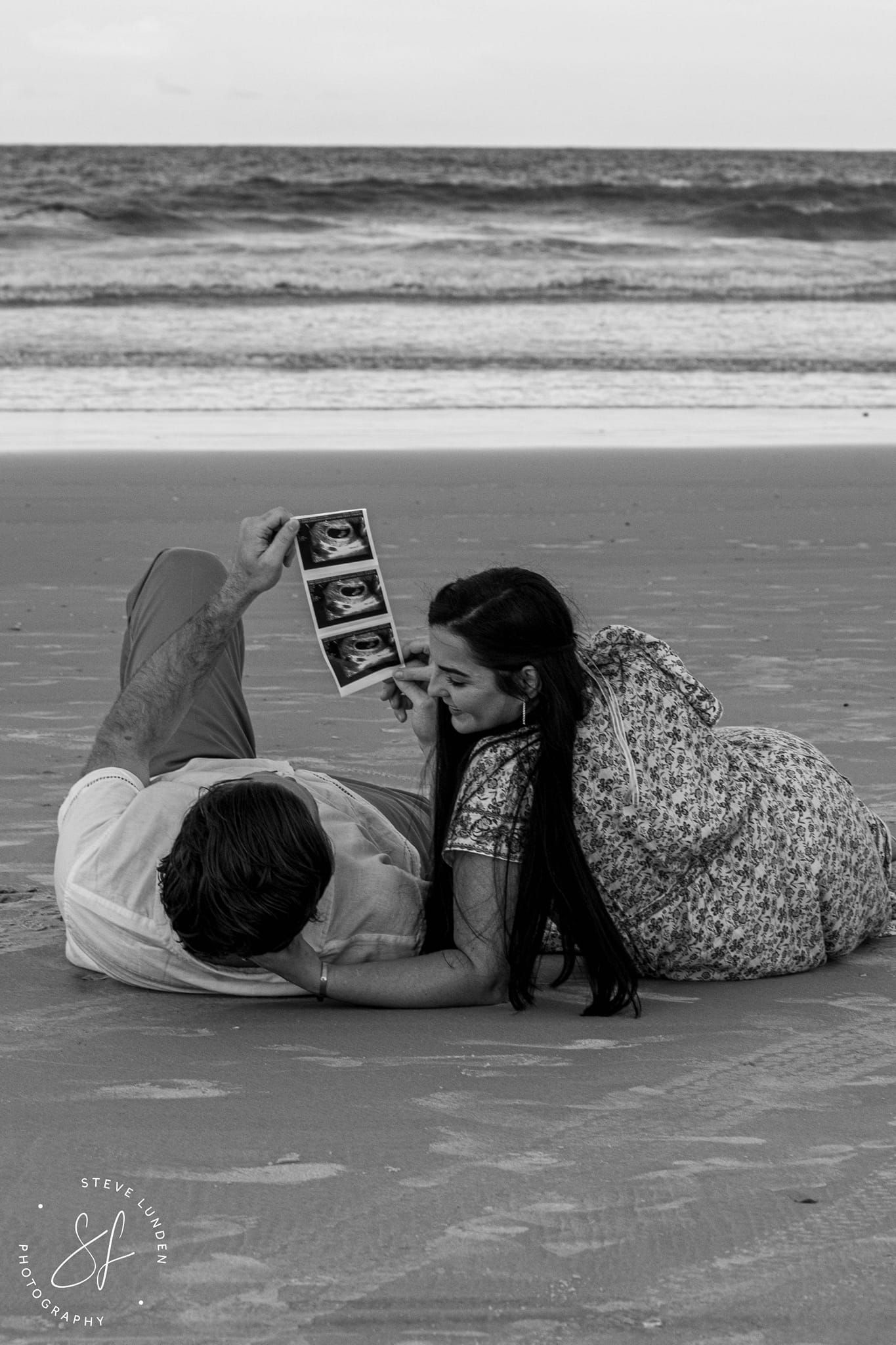 couple laying on beach looking at ultrasound of new baby