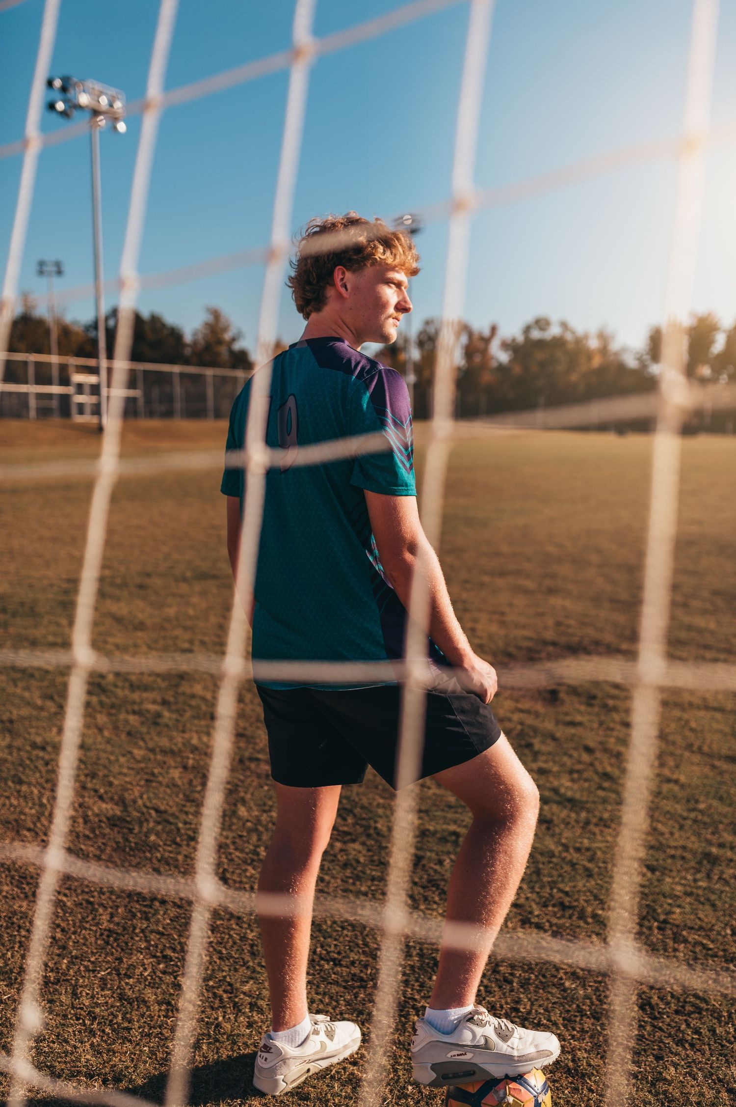 Athletic scene through soccer net showing player in navy uniform at sunset.
