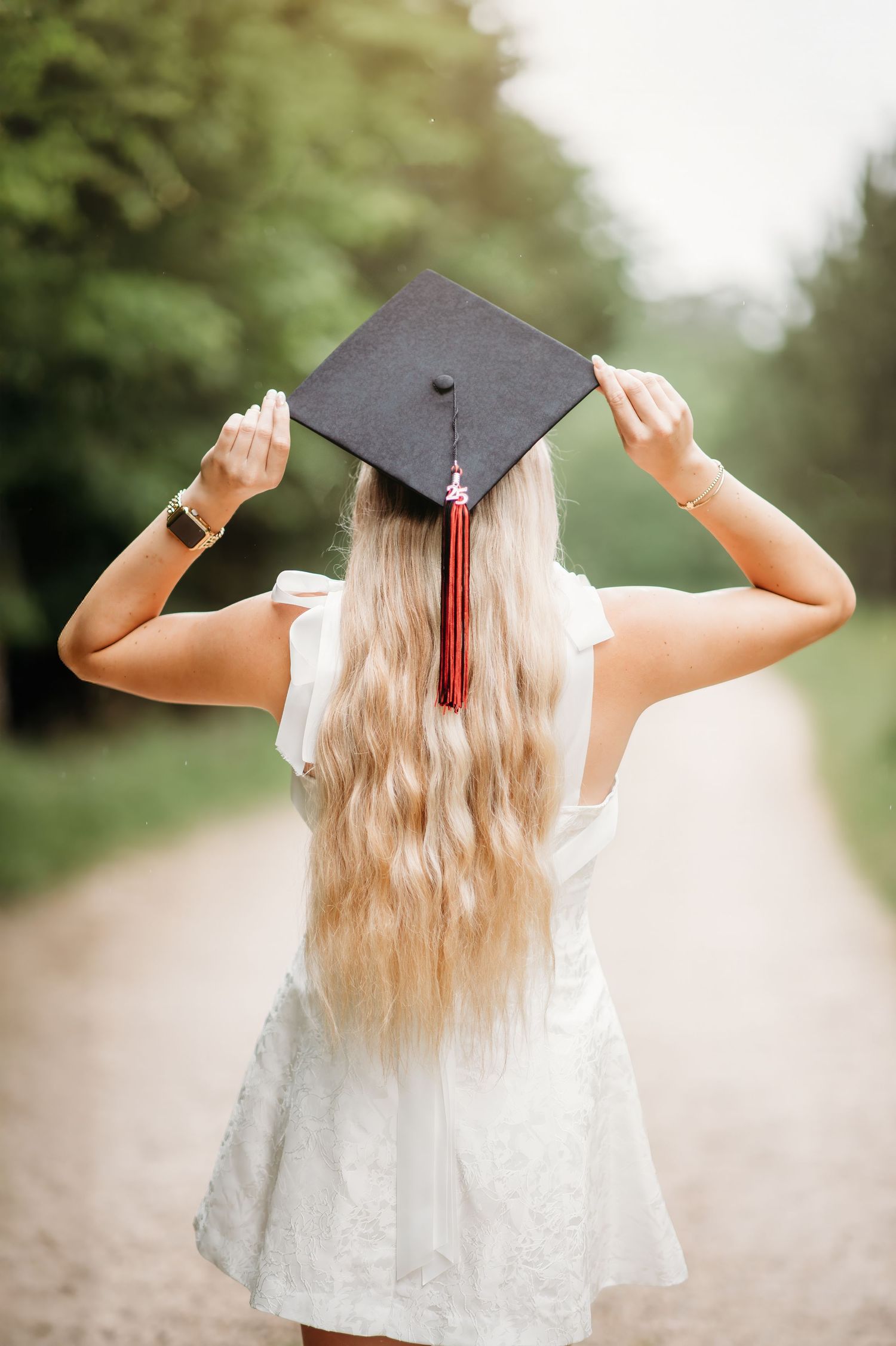 Graduate in white dress and cap celebrating with arms raised on rural path. fort mill, sc class of 2027