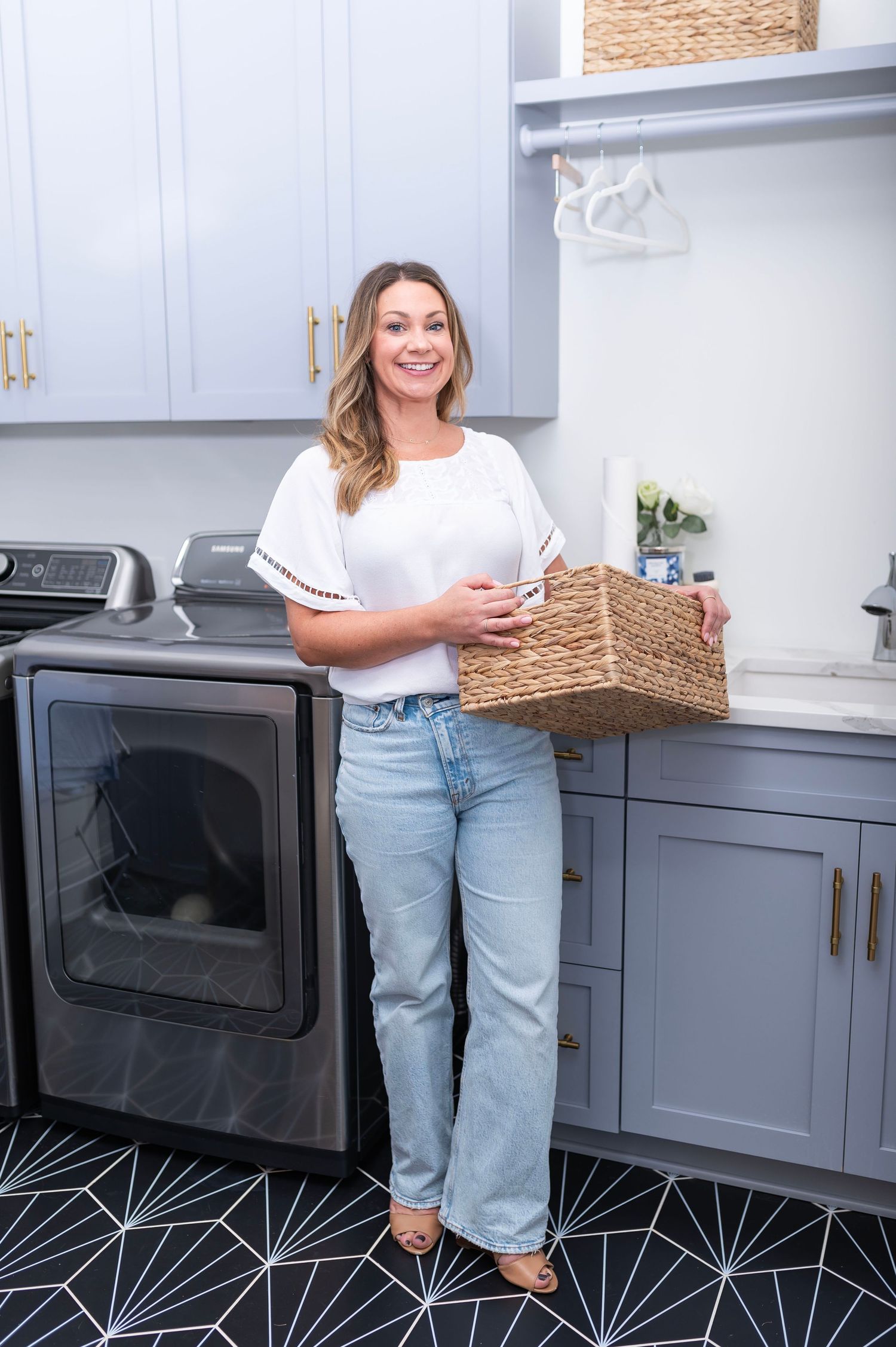 Person standing in a modern laundry room with gray cabinets and geometric floor tiles, holding a wicker basket.