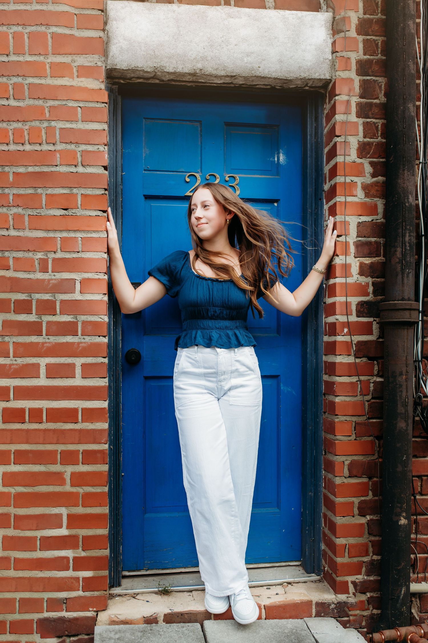 Person in blue top and white pants poses against bright blue door in brick doorway.