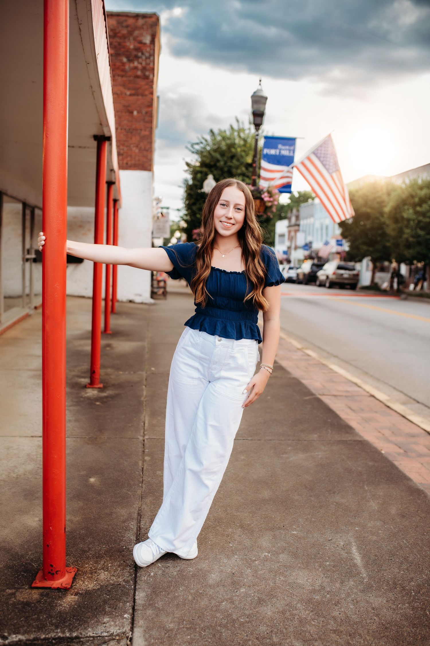 Person in navy top and white pants poses on sidewalk with American flag in background.