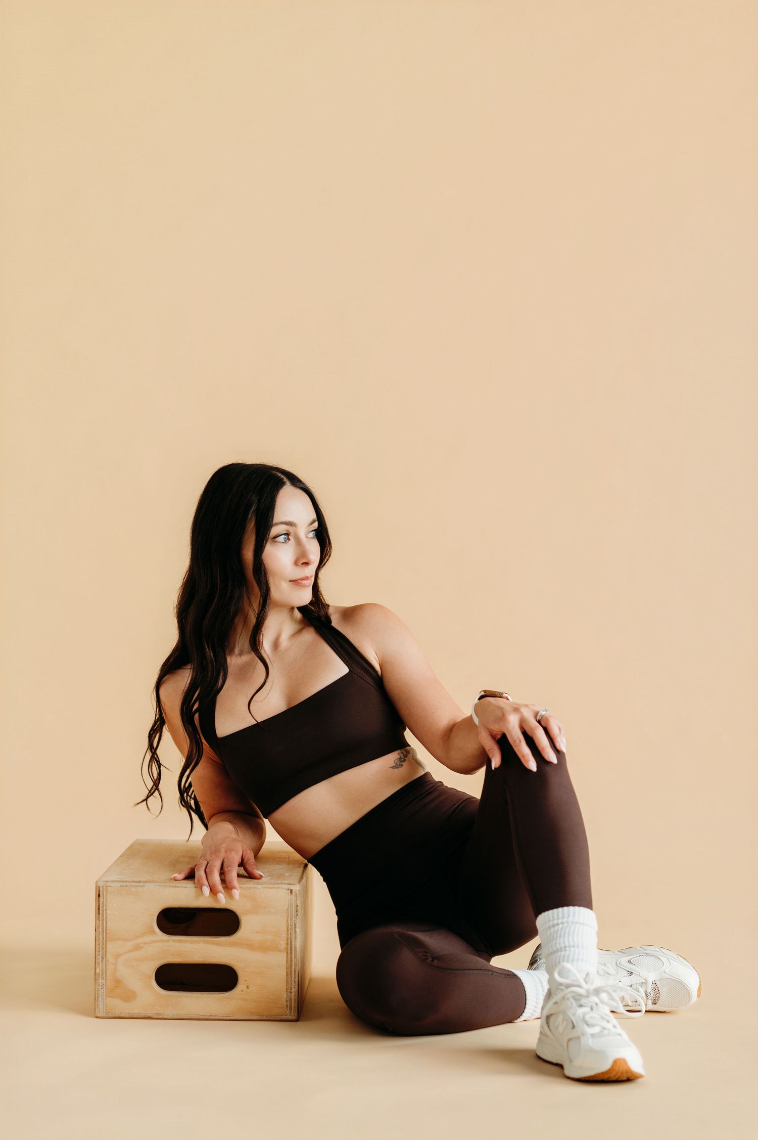 Woman in dark brown athletic wear sitting on wooden box against beige background, wearing white sneakers.