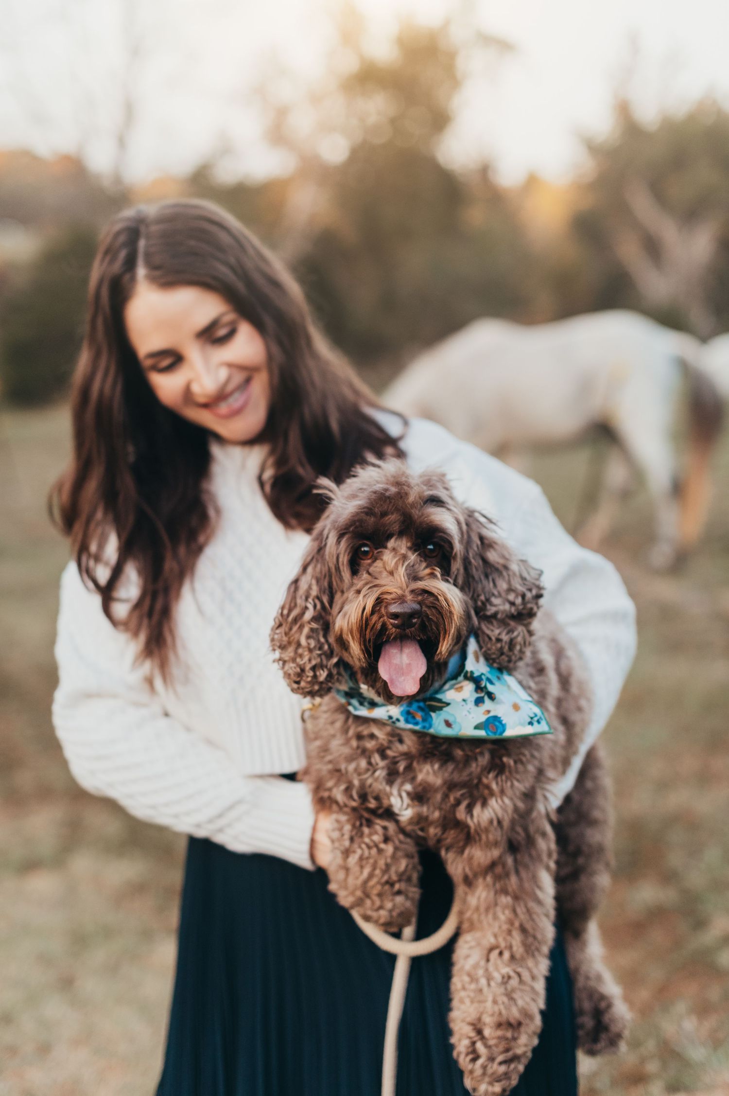 Person in white sweater holds smiling brown labradoodle dog wearing blue bandana in outdoor autumn setting.