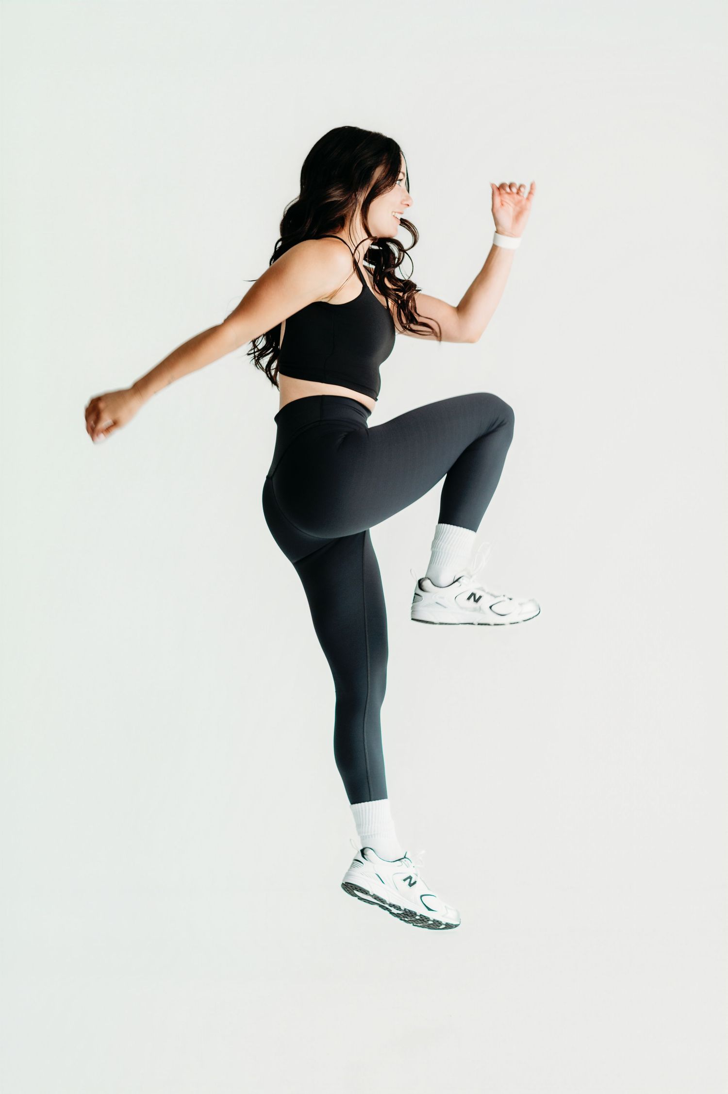 Woman in black athletic set and white sneakers jumping mid-air against white background.