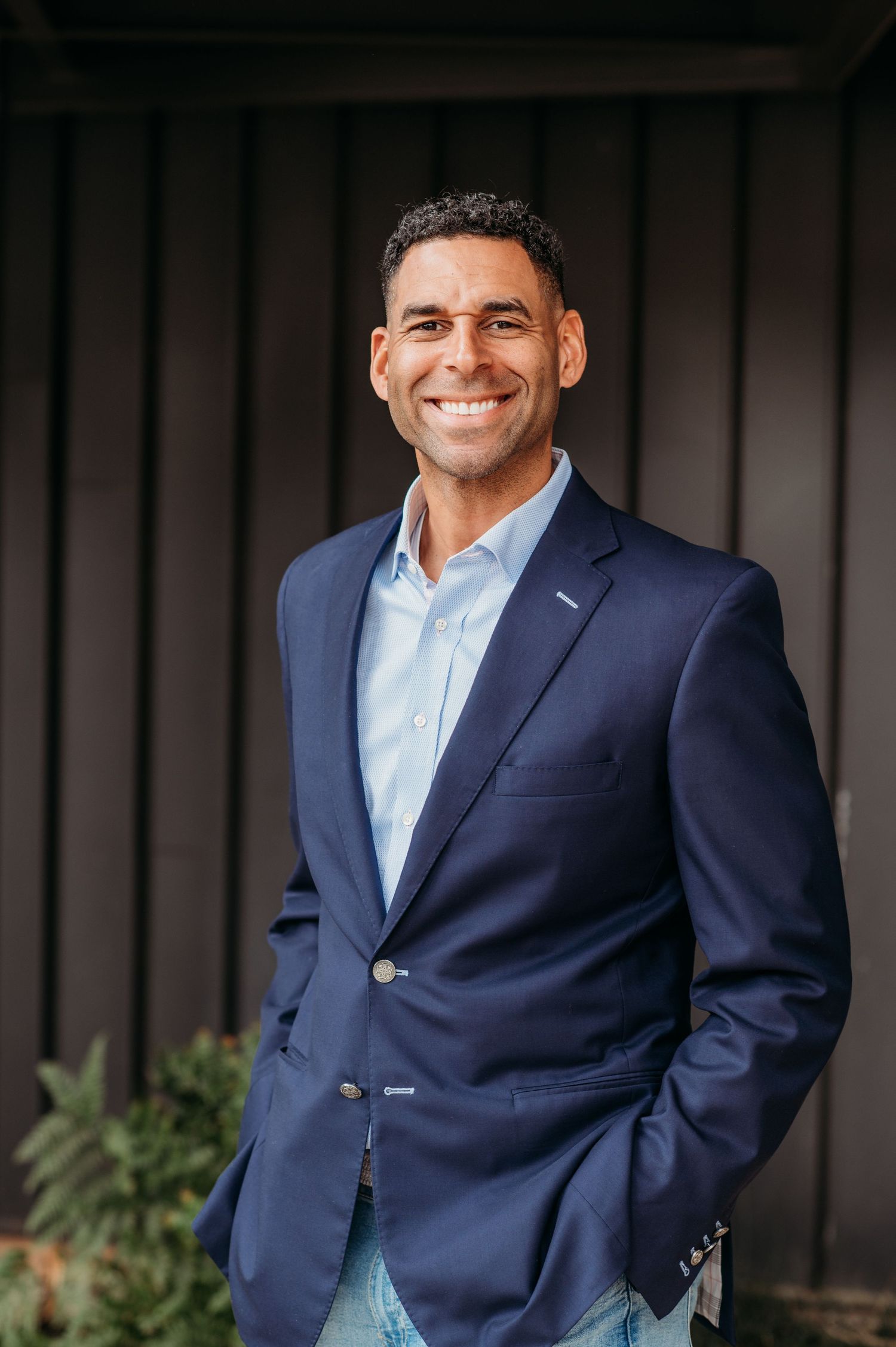 Professional headshot of a person wearing a navy blazer and light blue dress shirt smiling warmly against a dark background.