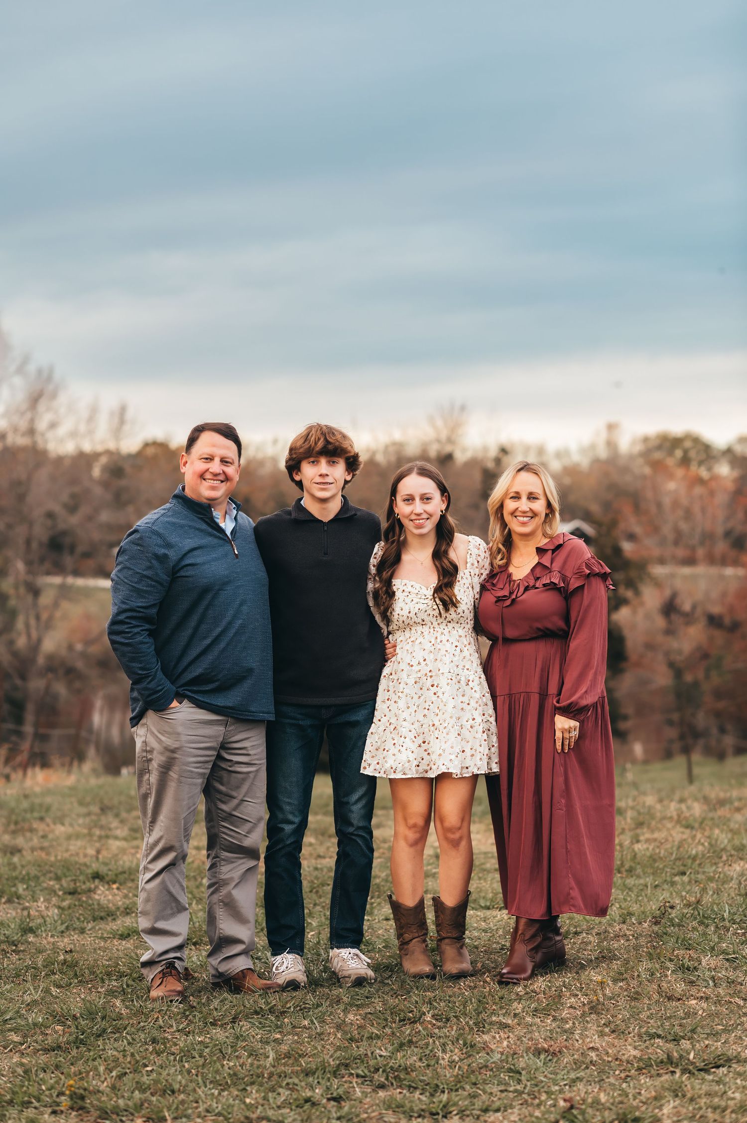 A family of four poses together outdoors on a grassy hill during autumn with overcast skies in the background.