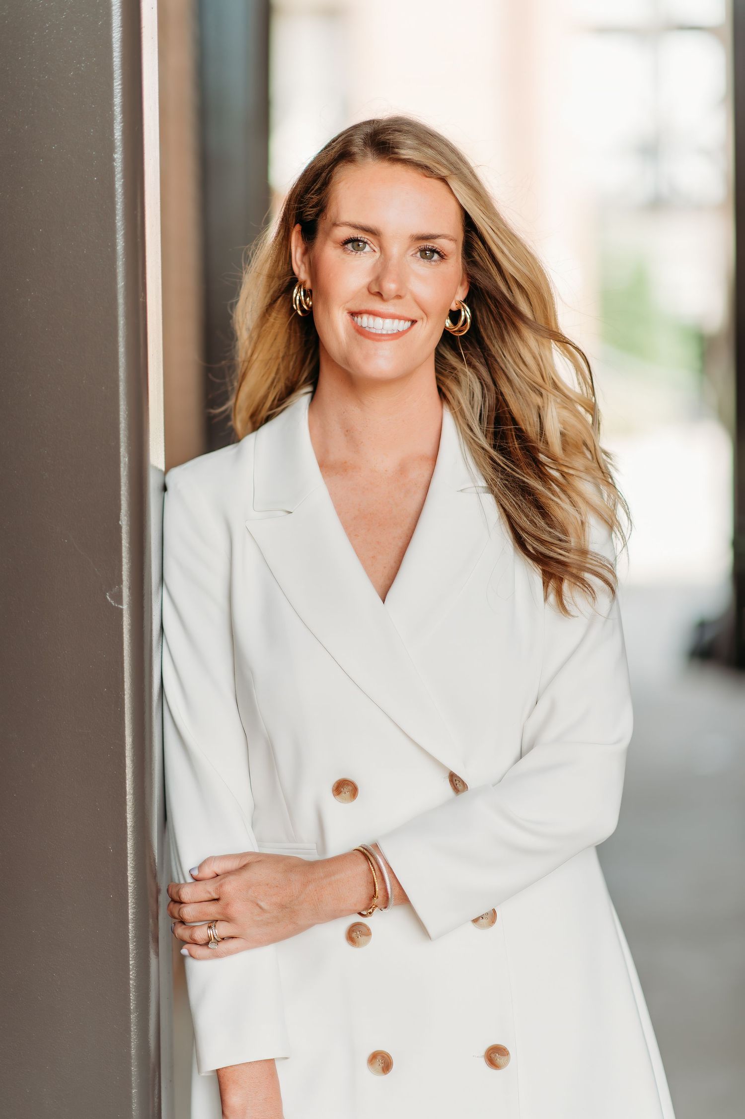 A professional wearing a white double-breasted blazer leans against a wall in an office setting with natural lighting.